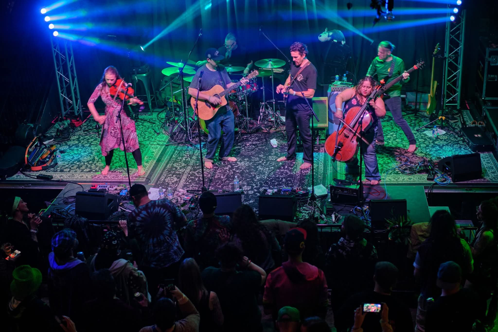 Musicians perform on stage with instruments, colorful lights, and an audience watching, confetti on the floor.