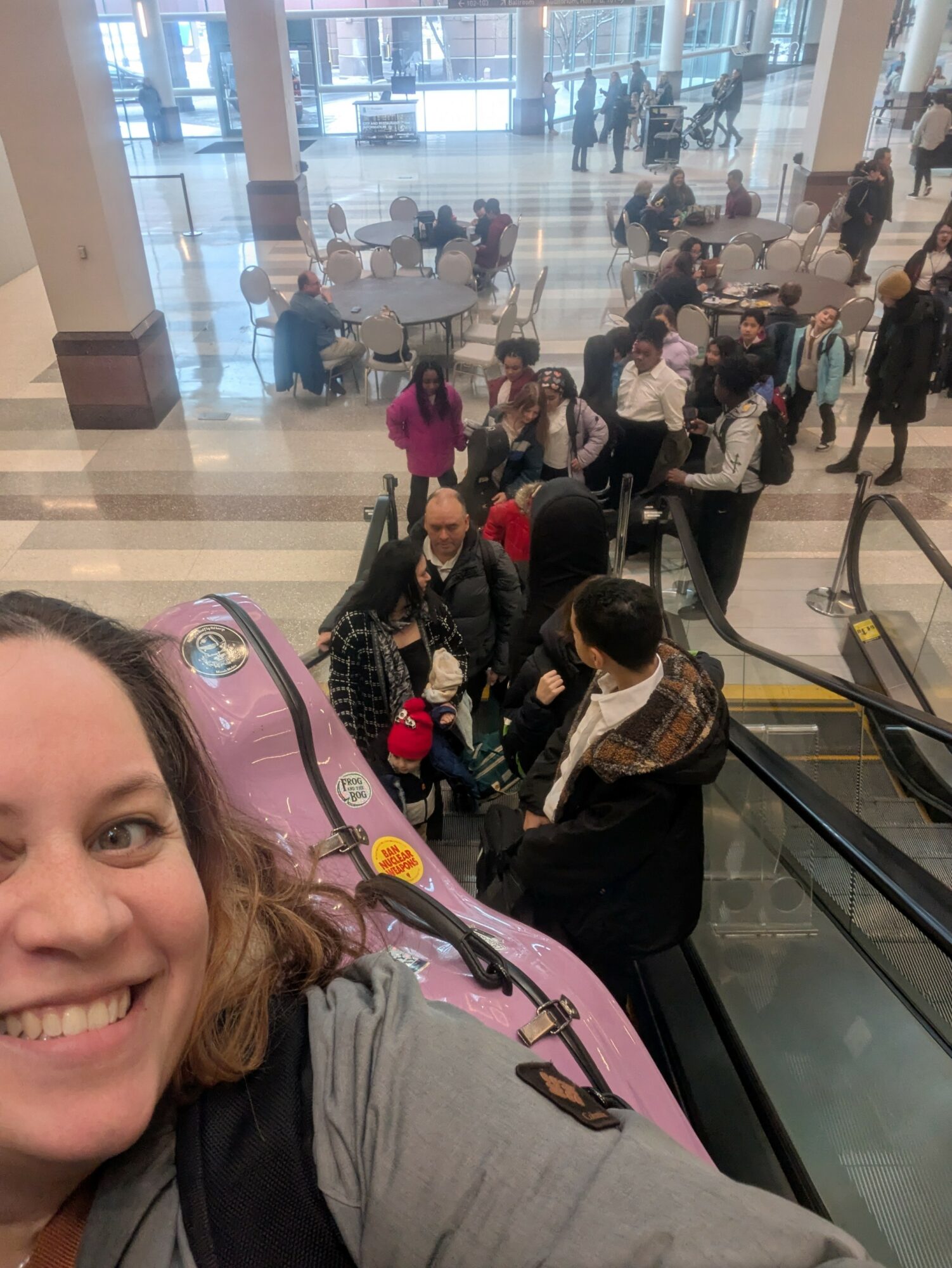 Woman taking selfie on escalator with people sitting and walking in background inside a building.