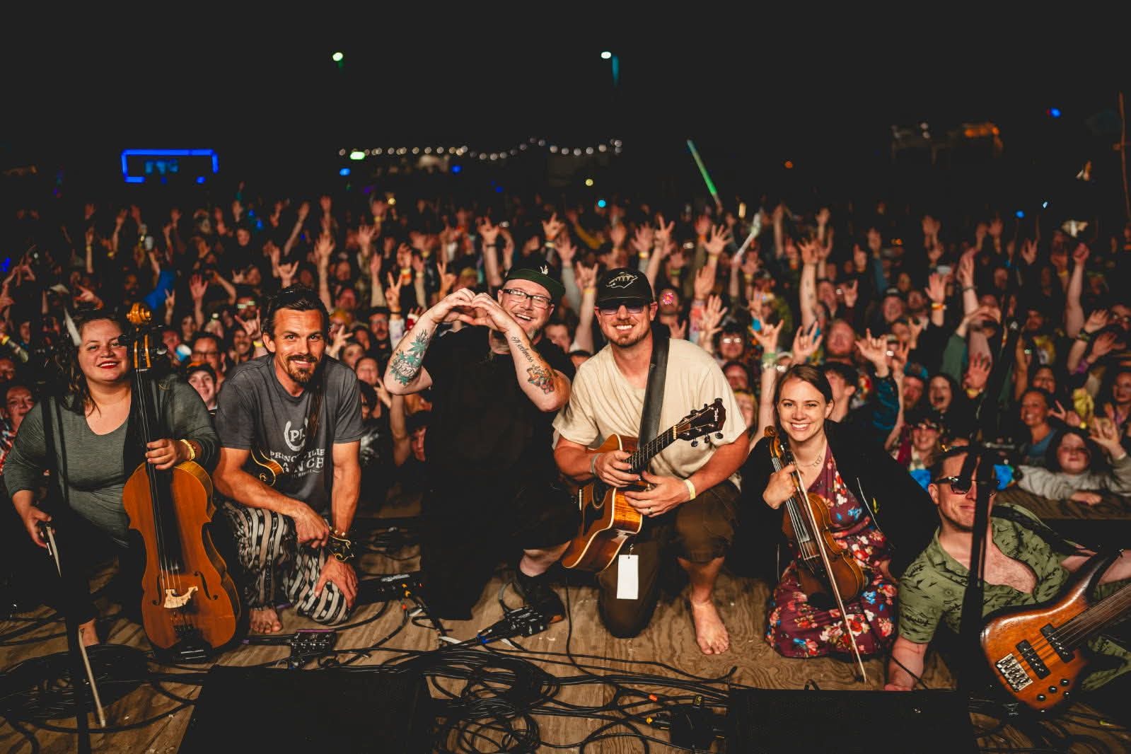 Band performs on stage with a large crowd in the background at night, some musicians holding instruments, smiling, and posing.