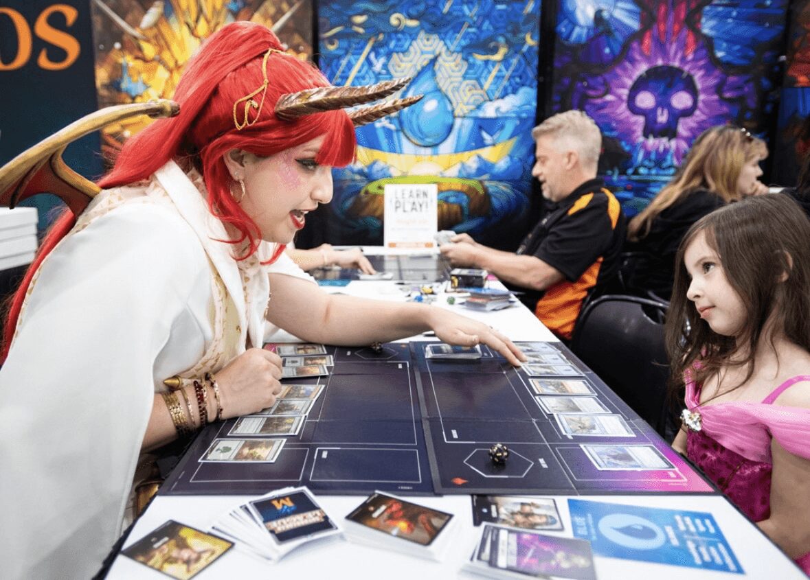 A woman with red hair and a white costume plays a tabletop game with a young girl at a gaming event, colorful background.