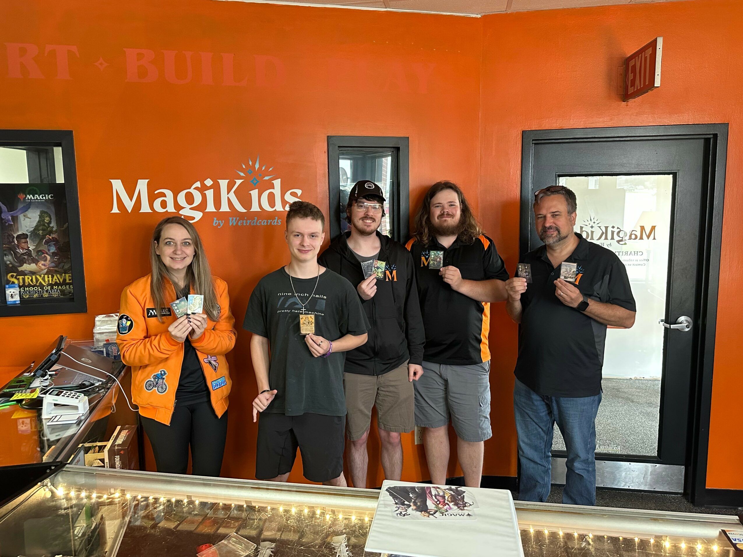 Five people stand in front of an orange wall with a sign that reads 'Magikids,' holding cards or tickets, smiling.