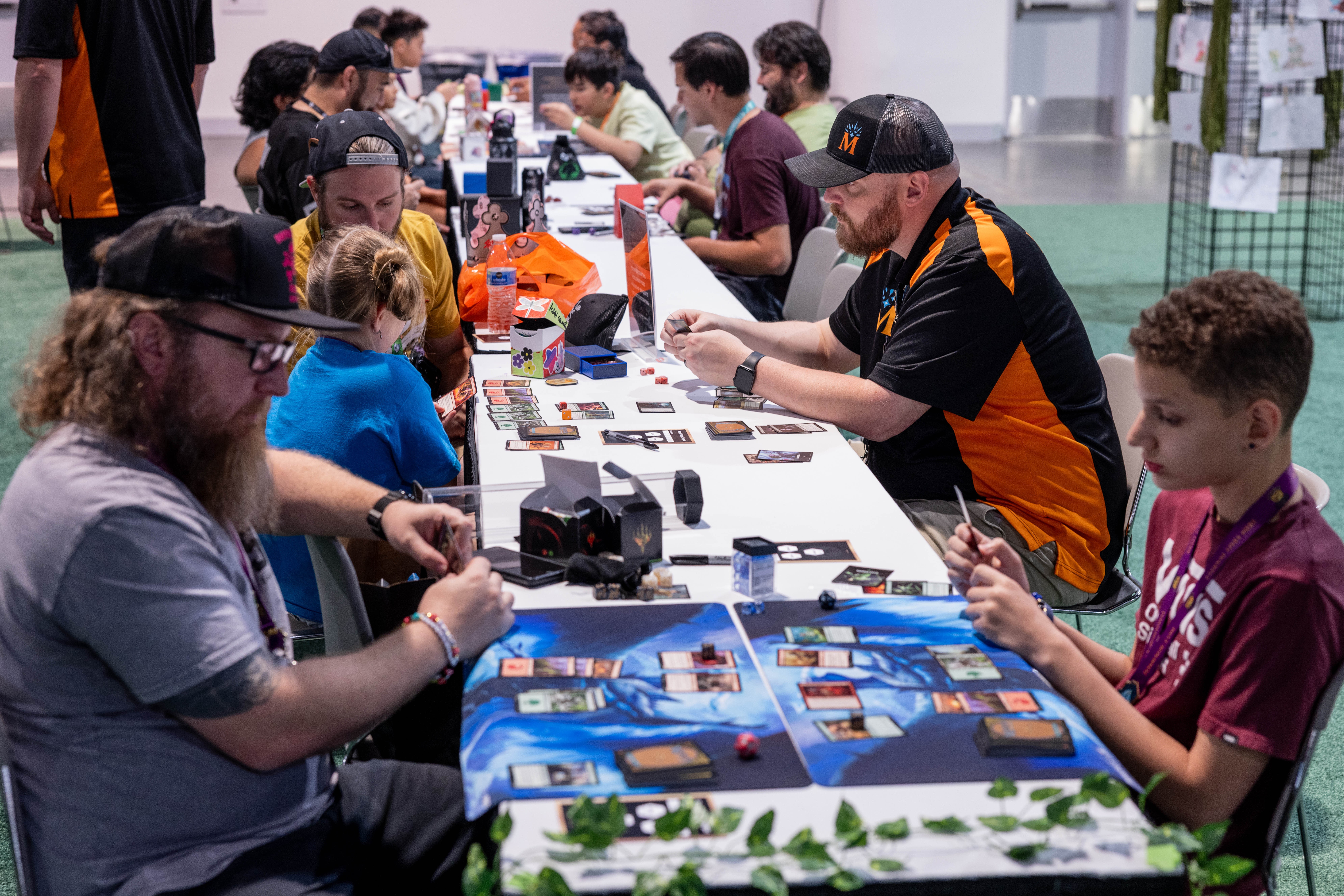 People playing a card game at a long table with game pieces and cards, in a bright indoor setting.