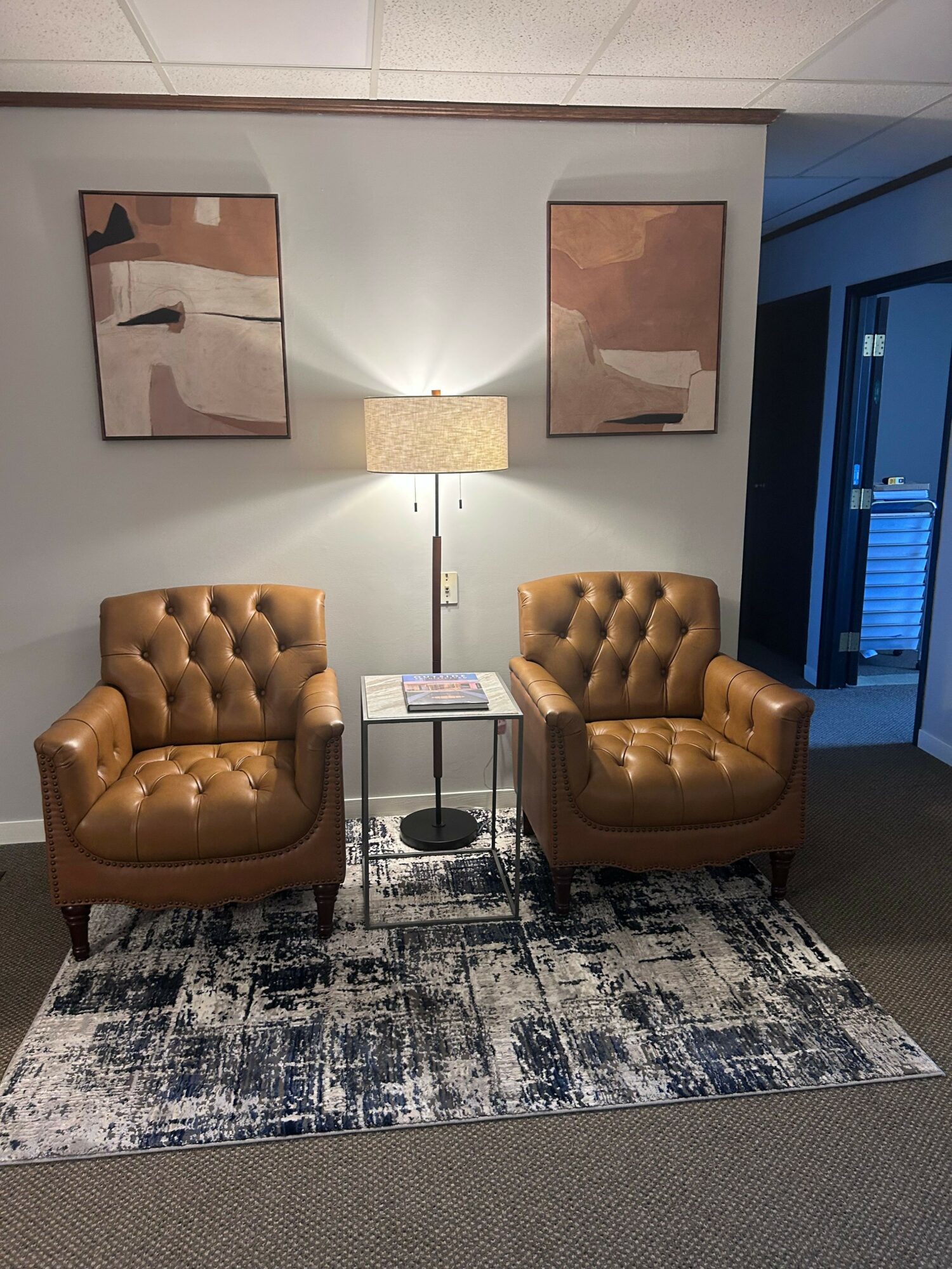Living room with two brown tufted armchairs, a small glass table, a patterned rug, and wall art, with a window in the background.