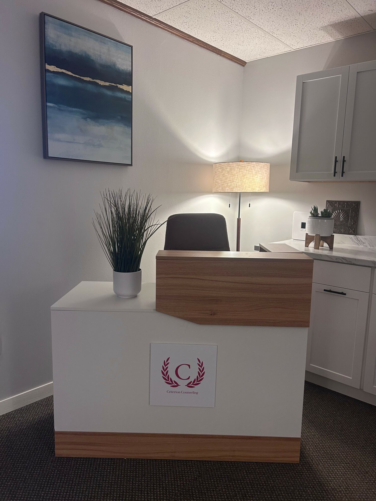 Office desk with a potted plant, a sign, and a wooden surface, with a kitchen area in the background.