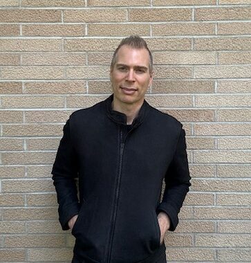 Man with short hair in a black jacket standing against a brick wall, hands in pockets.
