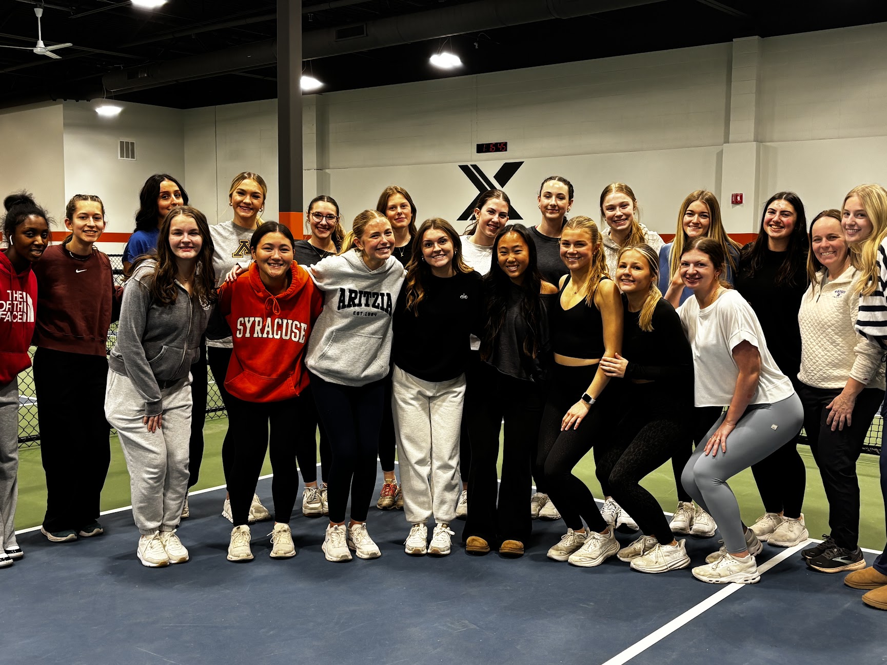 Group of people standing together indoors, some smiling, in a sports or training facility with a logo on the wall.