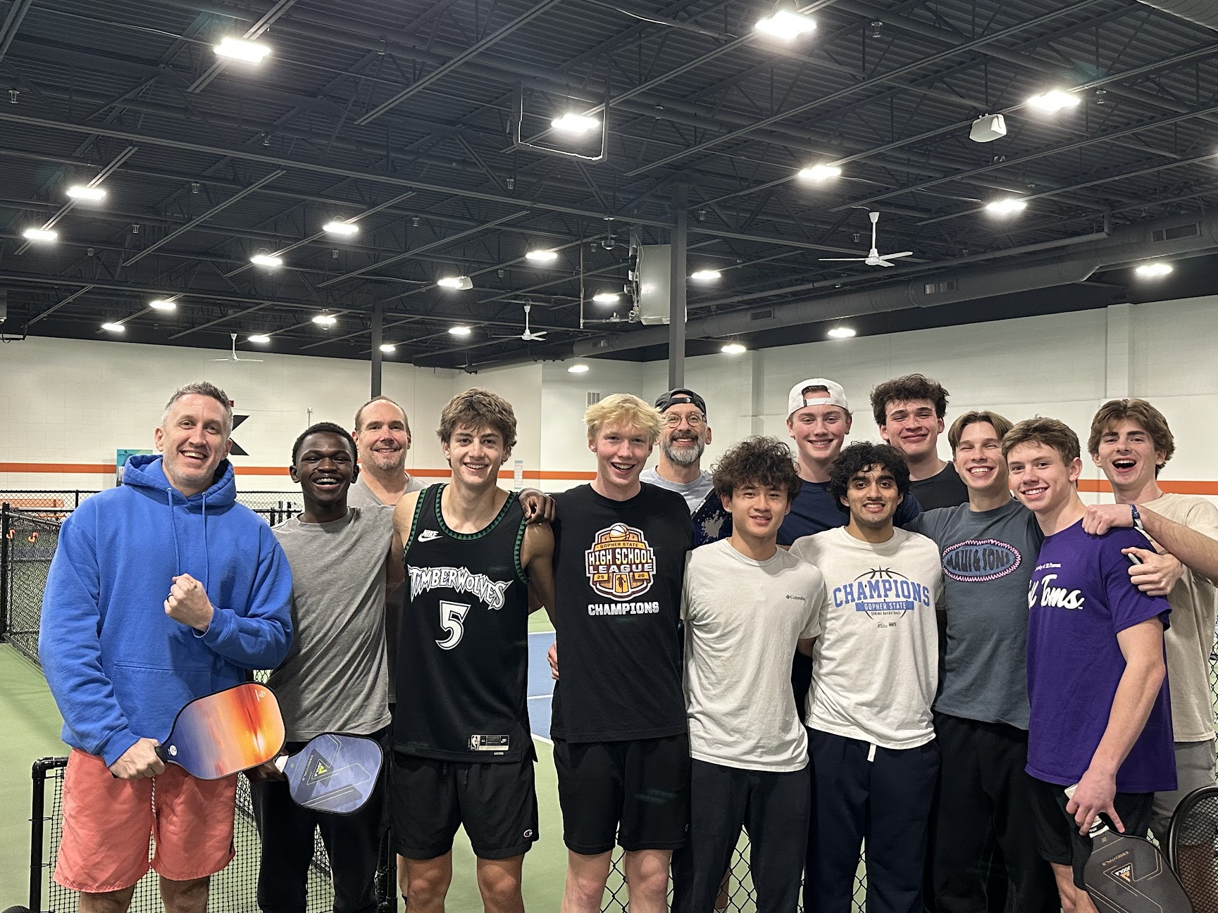 Group of young men standing together indoors, some smiling, wearing sports jerseys and casual clothing.