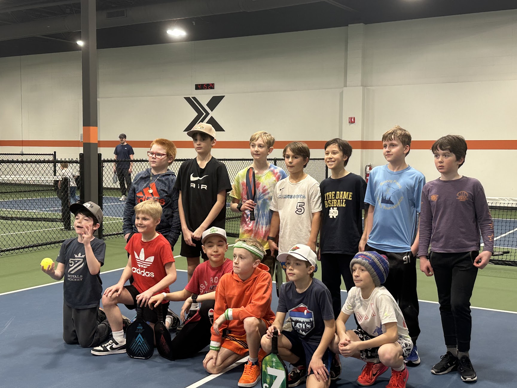 Group of children and teenagers posing on an indoor tennis court, some holding tennis balls, with a net and wall in background.
