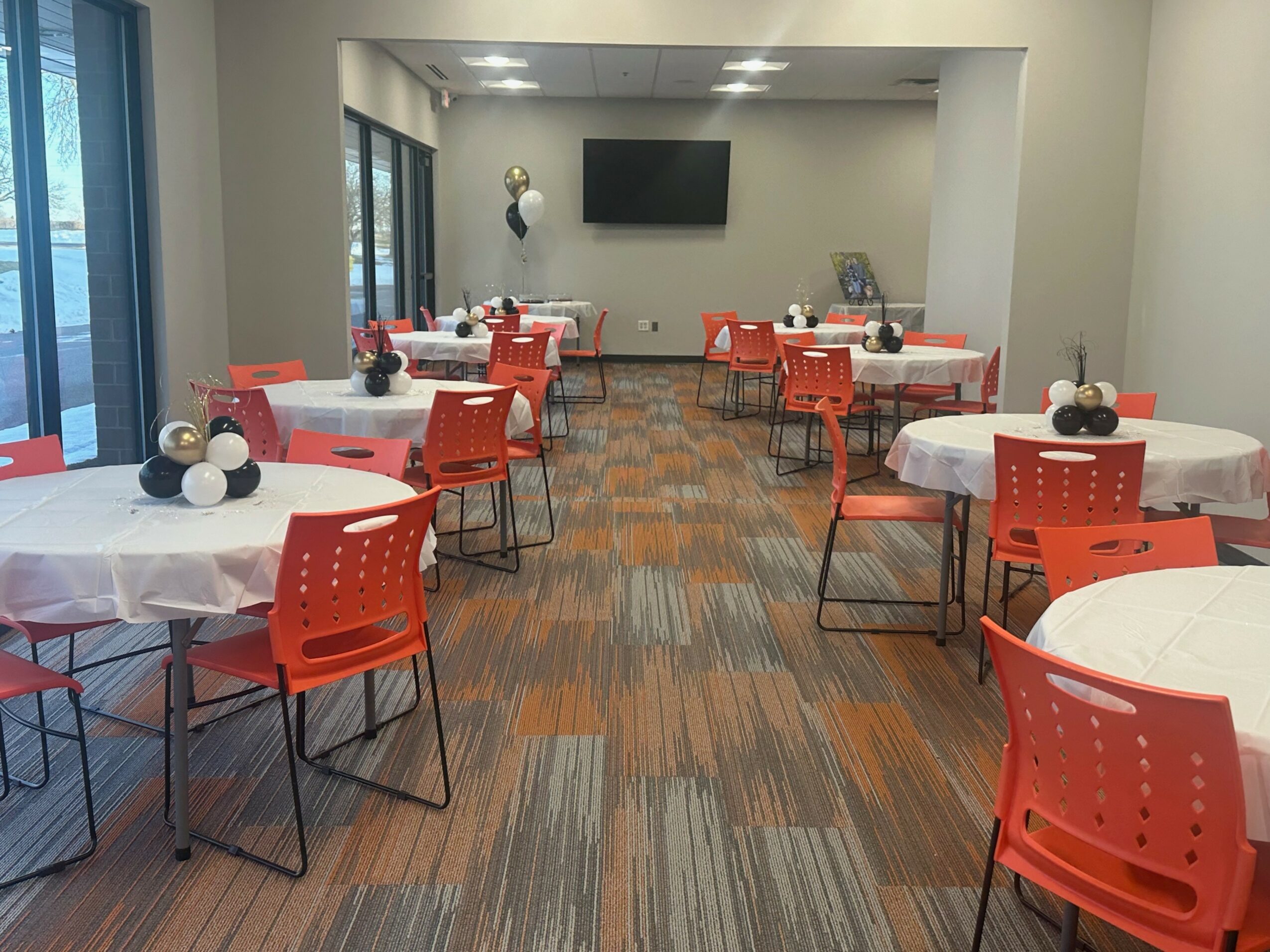Room with round tables covered in white cloths, orange chairs, and decorative centerpieces, with a TV on the wall and windows on the left.