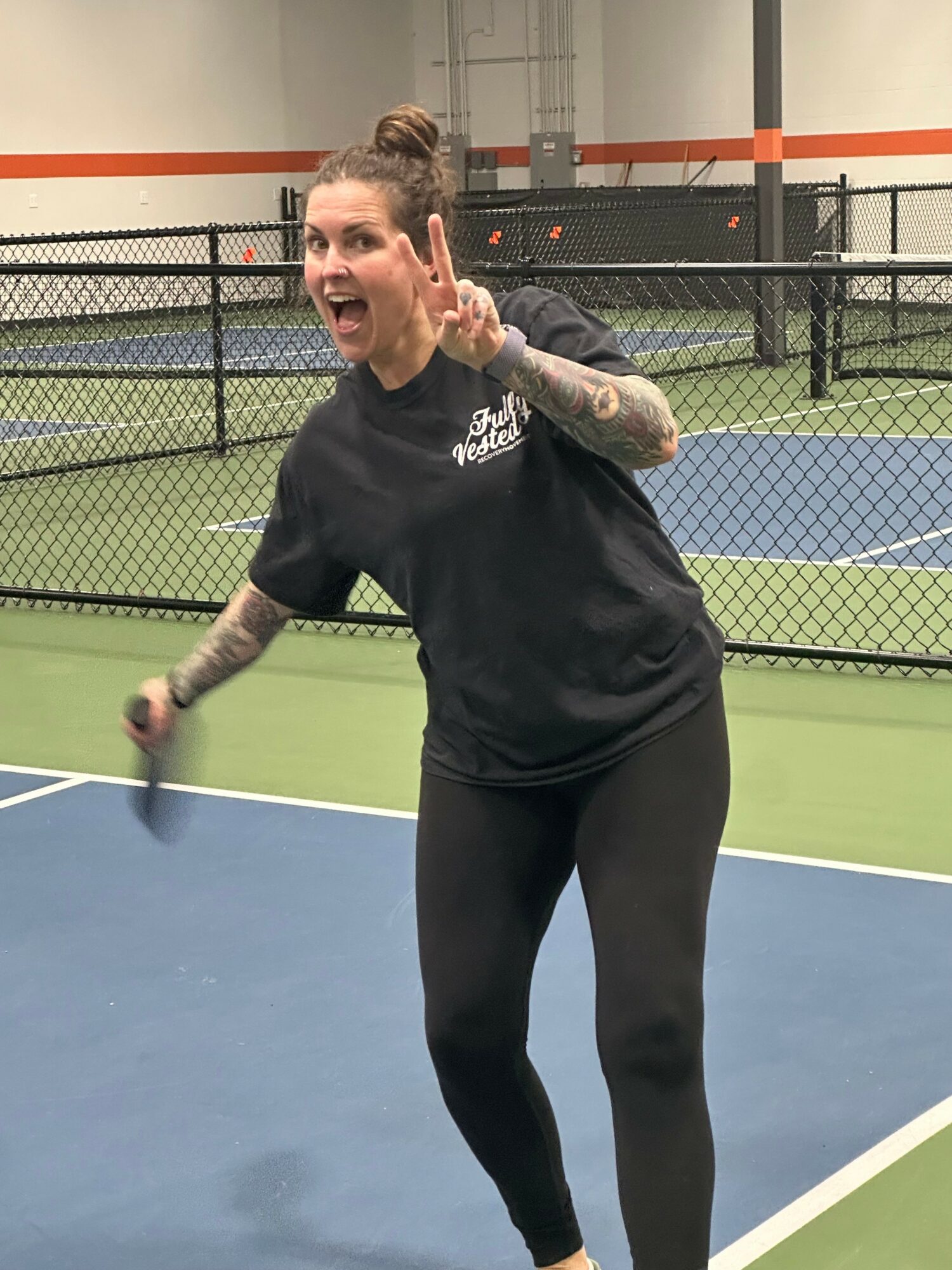 Woman with tattoos on arms smiling and making a peace sign at an indoor tennis court.