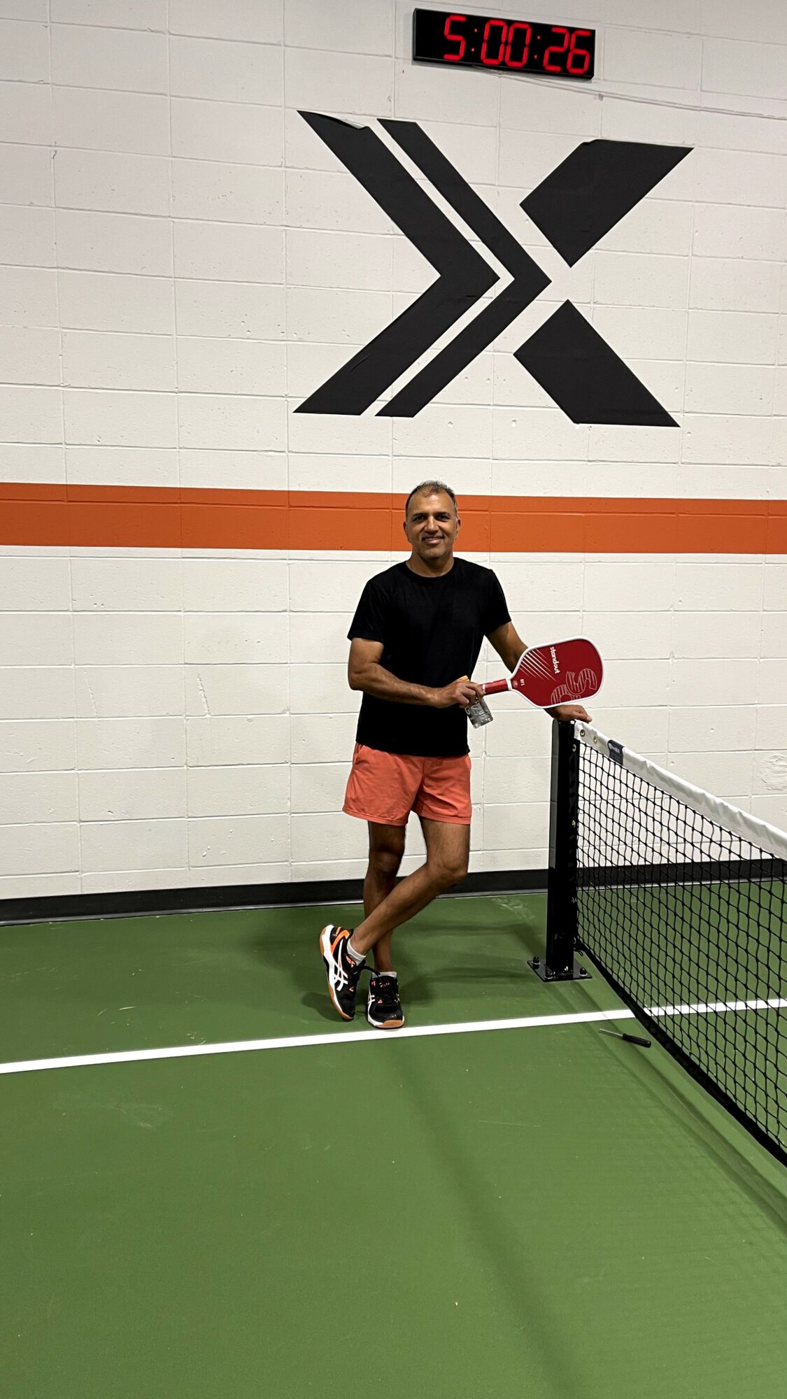 Person holding a paddle on a ping pong court with a large black and white logo on the wall behind.