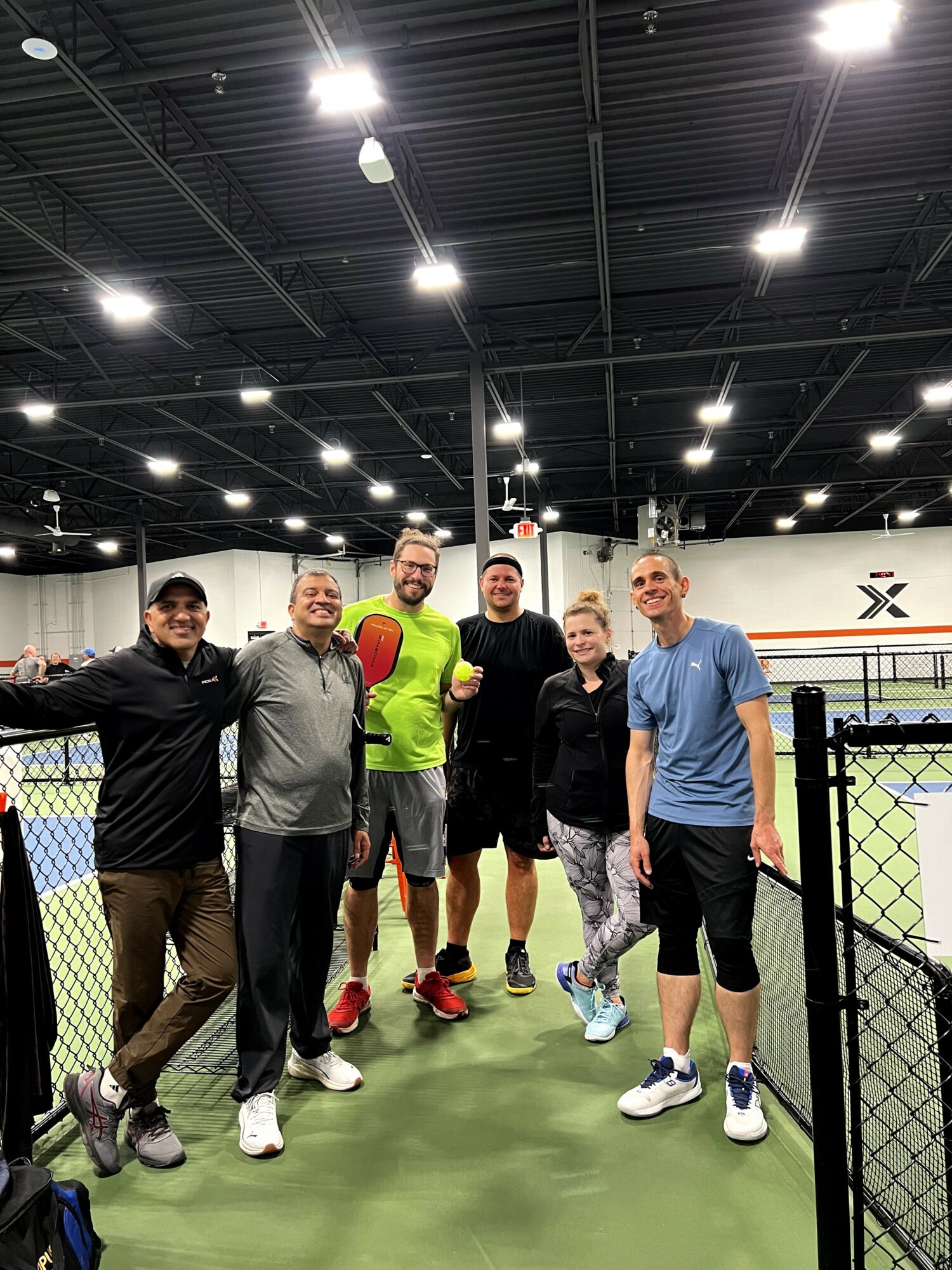 Group of six people standing on an indoor tennis court, smiling, with a high ceiling and bright lights above.