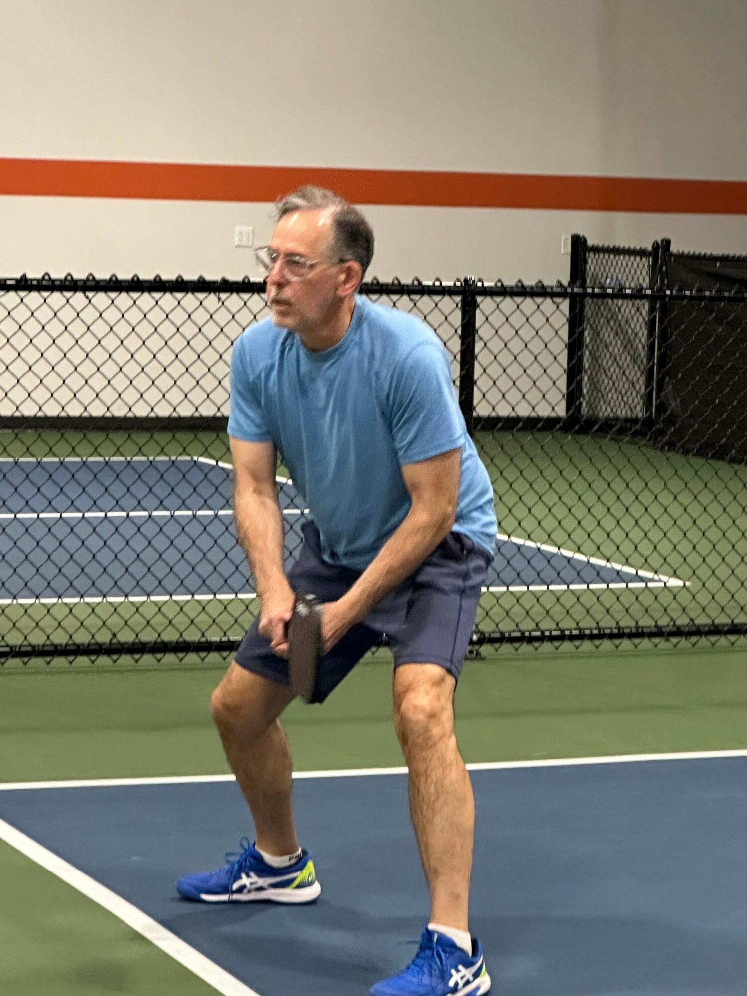 Man in blue shirt and shorts playing tennis on court, holding racket, in an indoor facility.