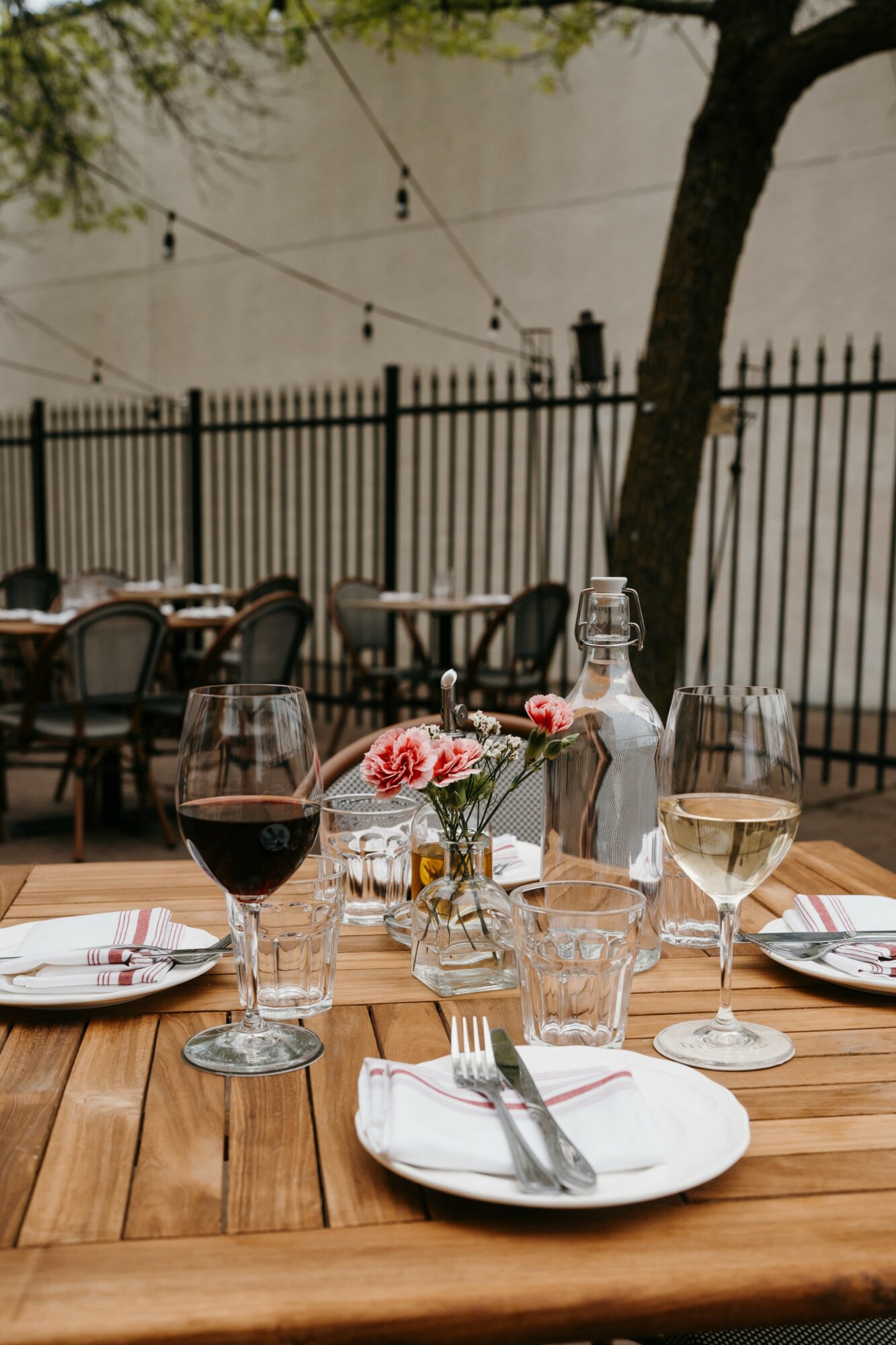 Outdoor dining table set with wine glasses, plates, cutlery, and a small flower arrangement, with a fence and trees in background.