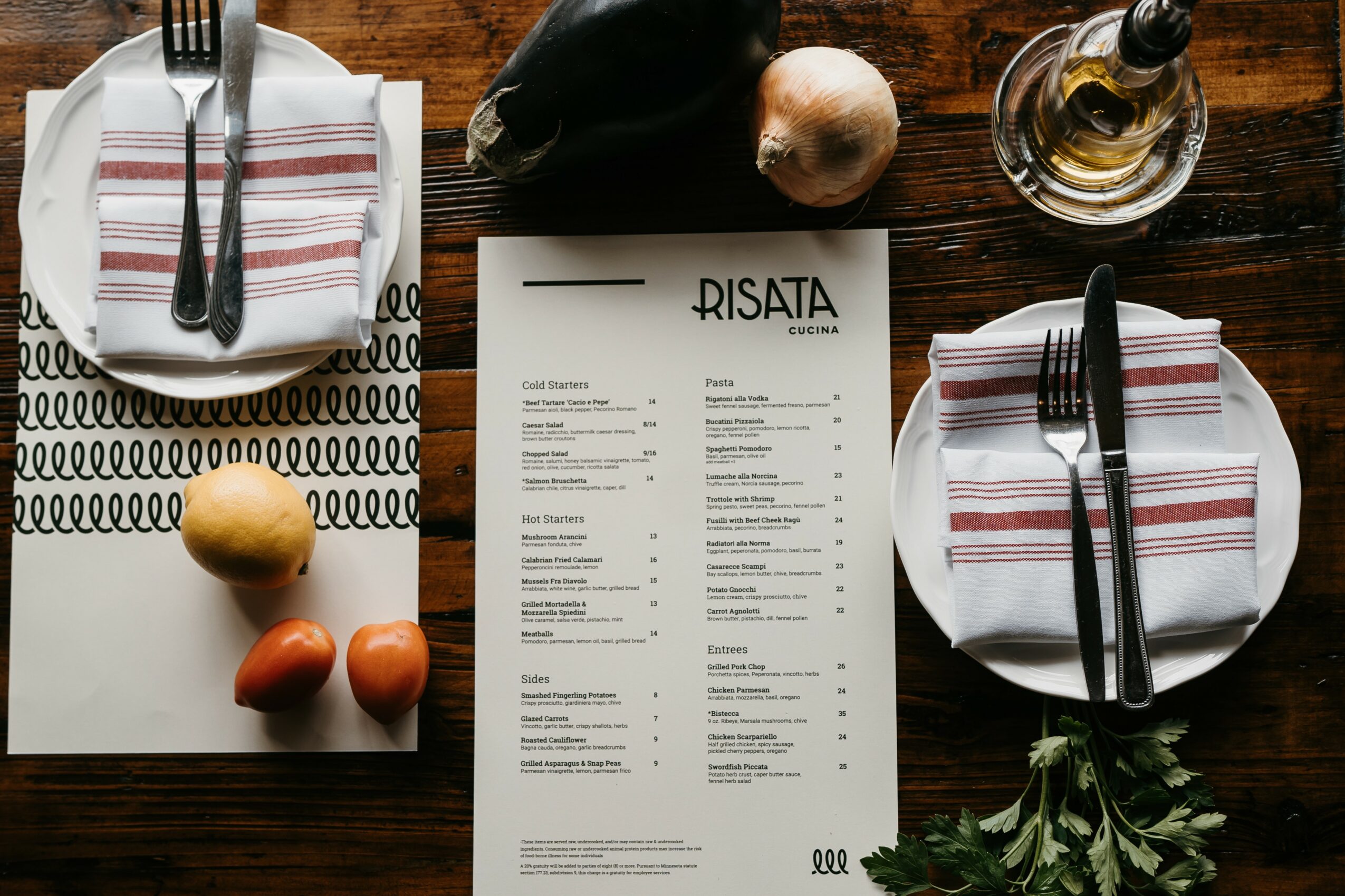 Table setting with plates, cutlery, a menu, onions, a bottle, and herbs on a wooden table.