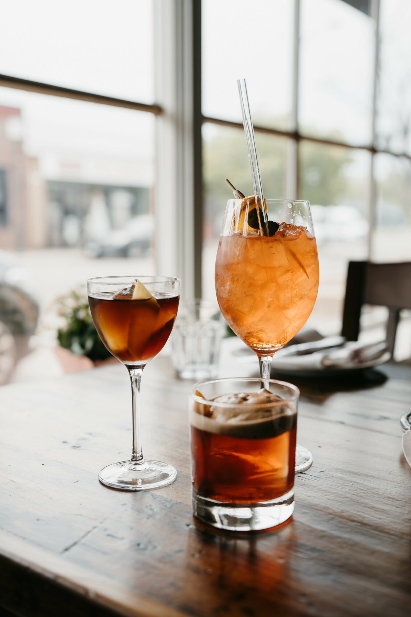 Three drinks on a wooden table near a large window, with a city view outside, including a wine glass, a cocktail, and a highball glass.
