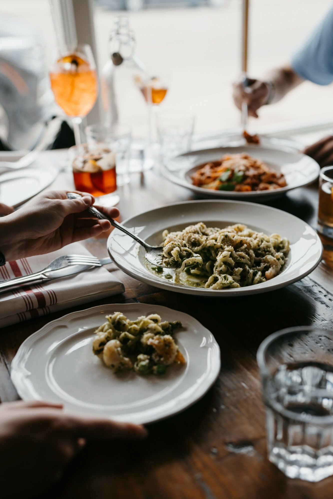 People dining with plates of pasta and drinks on a wooden table, hands reaching for food, glasses of orange beverage, and water.