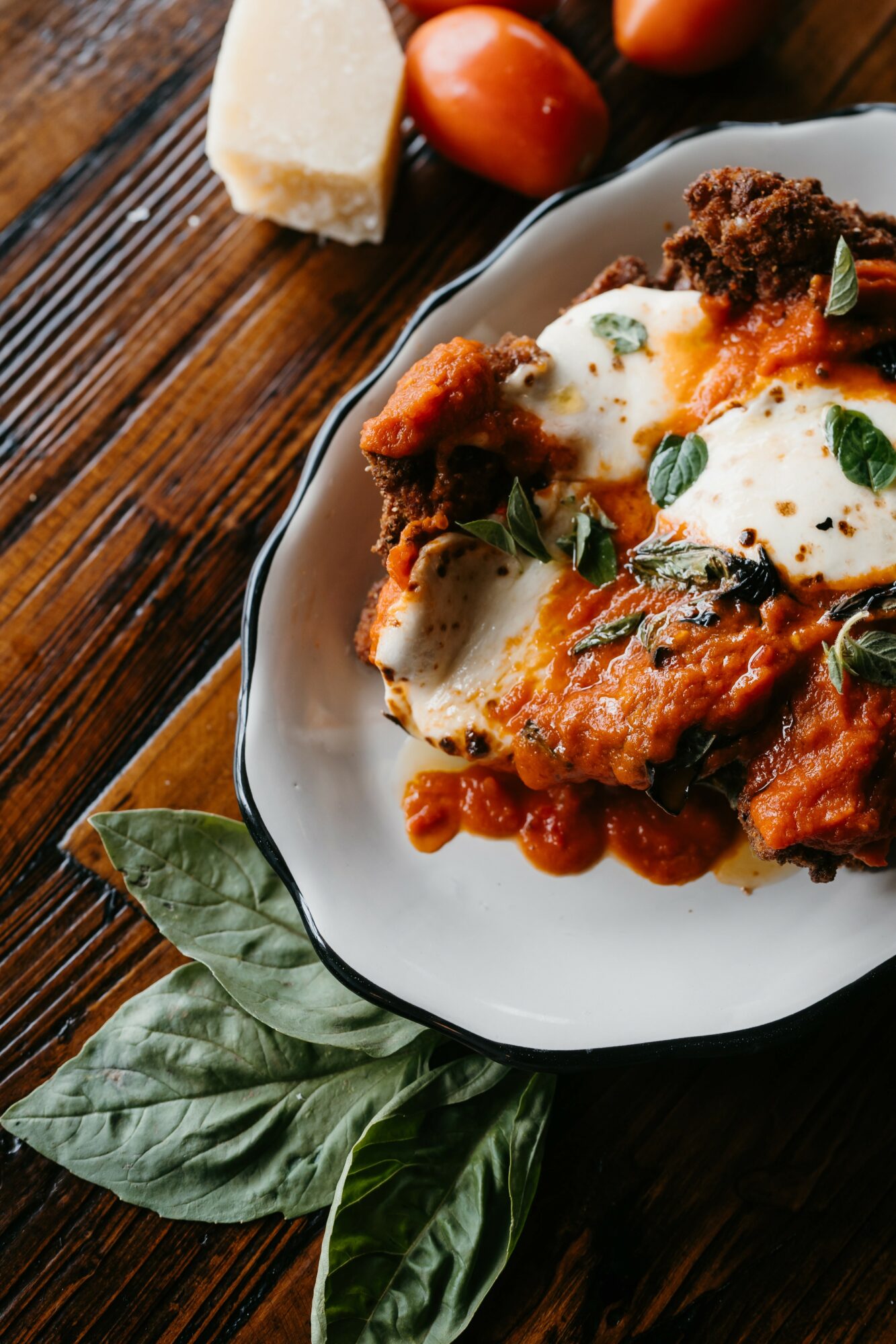 Plate of baked pasta with tomato sauce, mozzarella, and basil on a dark wooden table, with cherry tomatoes and cheese nearby.