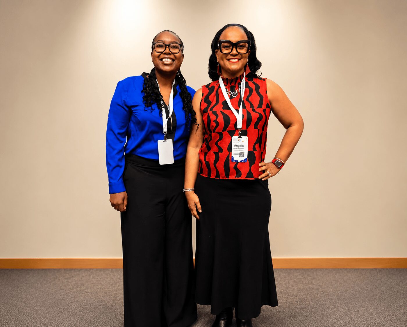 Two women stand side by side, smiling, wearing conference badges, one in a blue blazer and the other in a red patterned top.