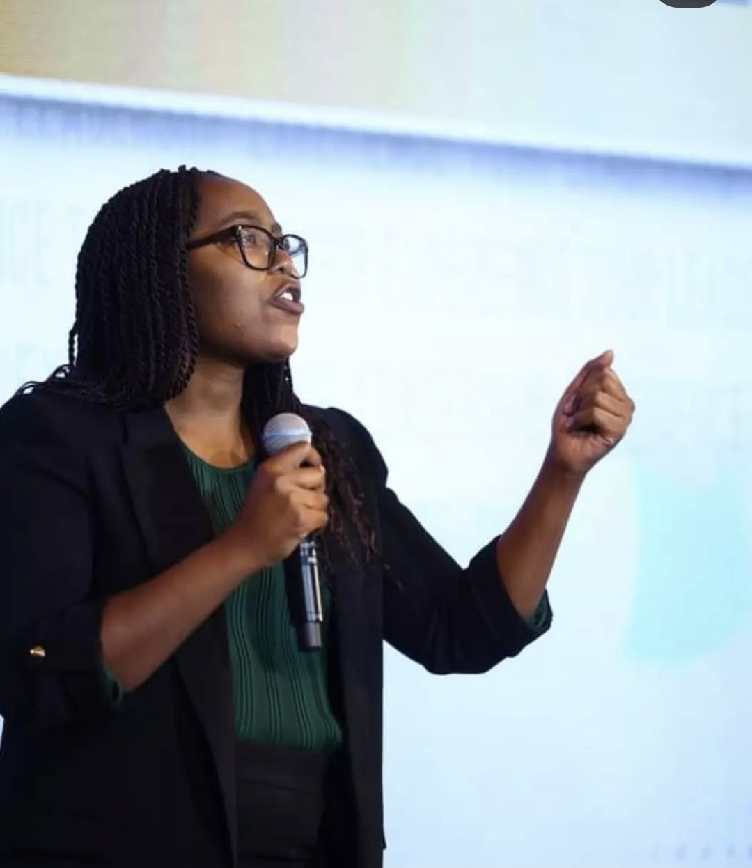 Woman with glasses speaking into a microphone, gesturing with her right hand, against a light background.