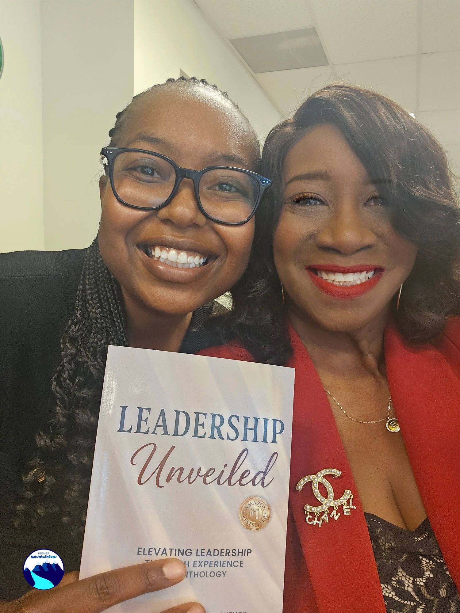 Two women smiling, holding a sign that reads 'Leadership Unveiled,' in an indoor setting with ceiling tiles visible.