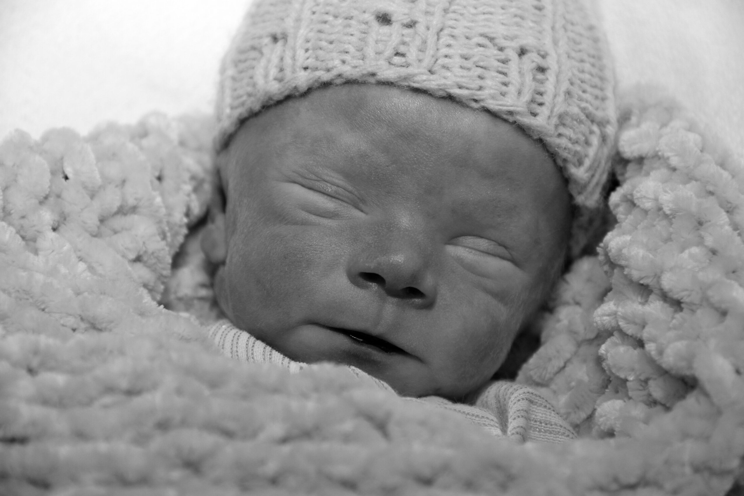 Newborn baby with closed eyes, wearing a knit hat, wrapped in a soft blanket.