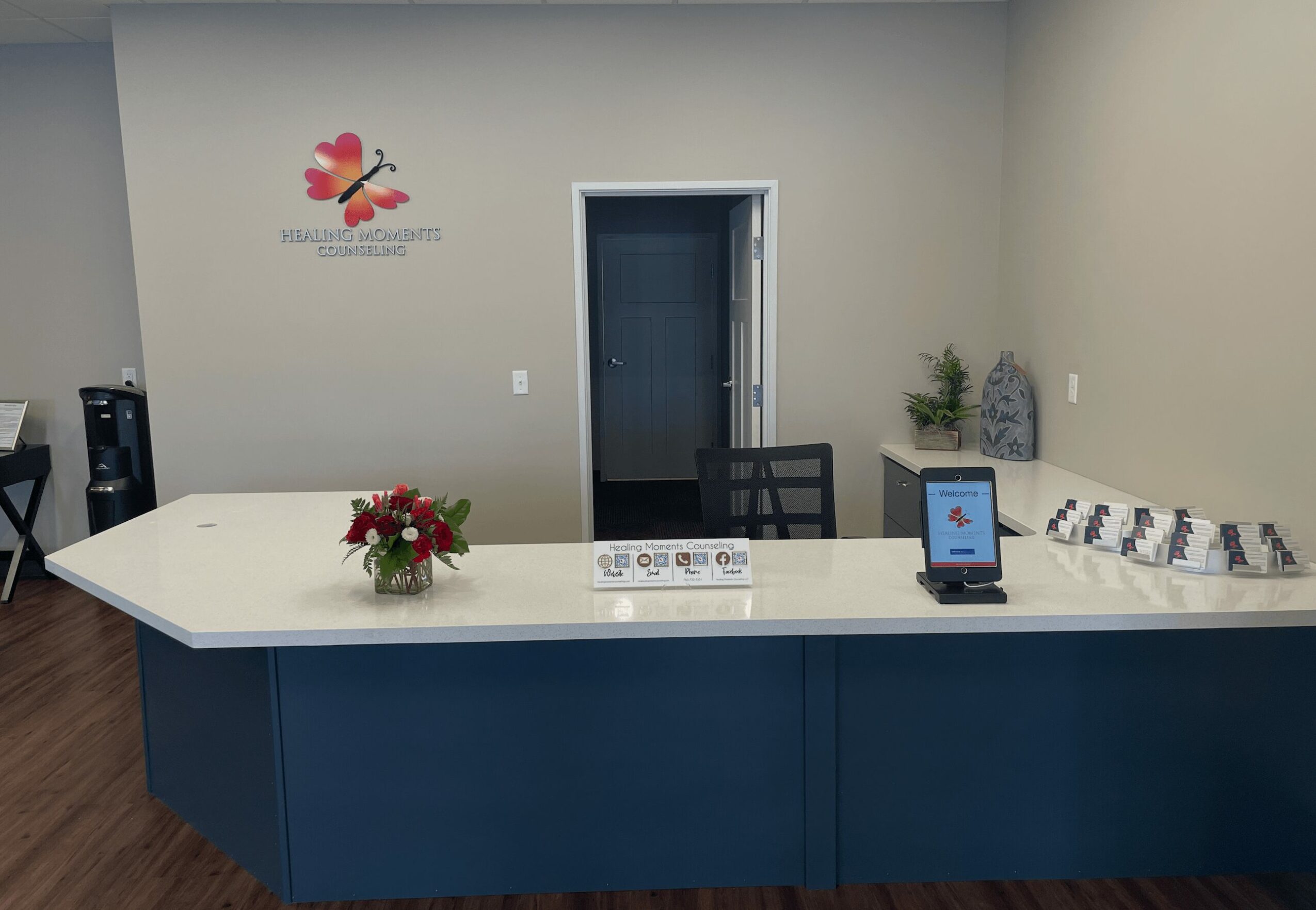 Reception desk with a flower arrangement, a tablet, and a row of pillows, in a room with a door and a logo on the wall.