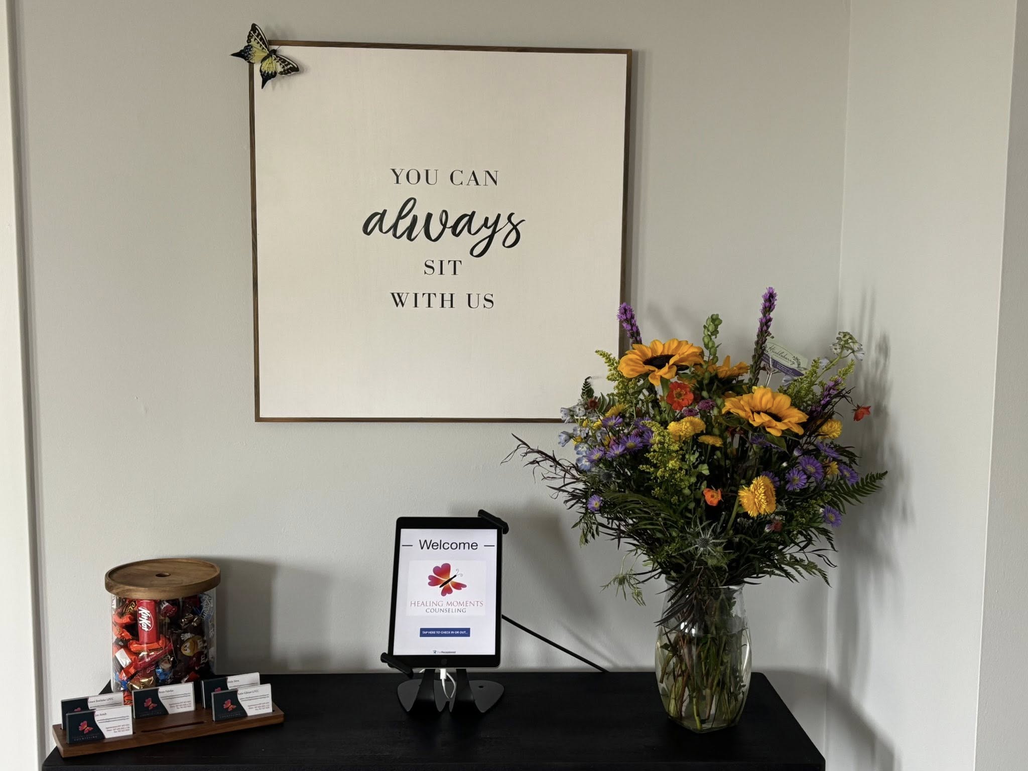 Wall with framed quote, flower arrangement in glass vase, and a small digital display on a table.