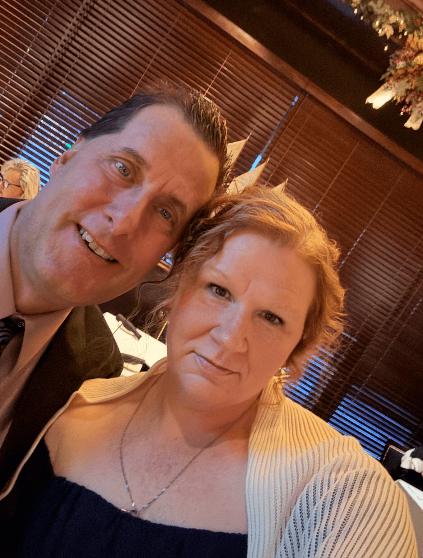 Two people smiling indoors with wooden blinds and floral decorations in the background.