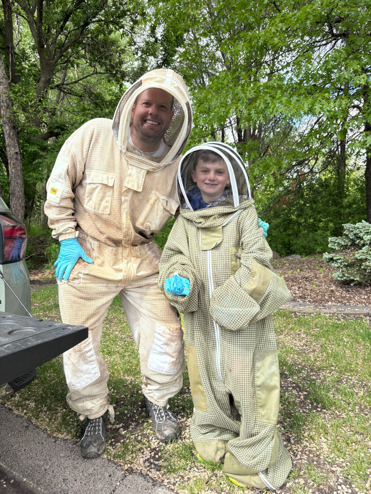 Two people in protective suits and helmets standing outdoors among trees, one holding a clipboard, in a natural setting.