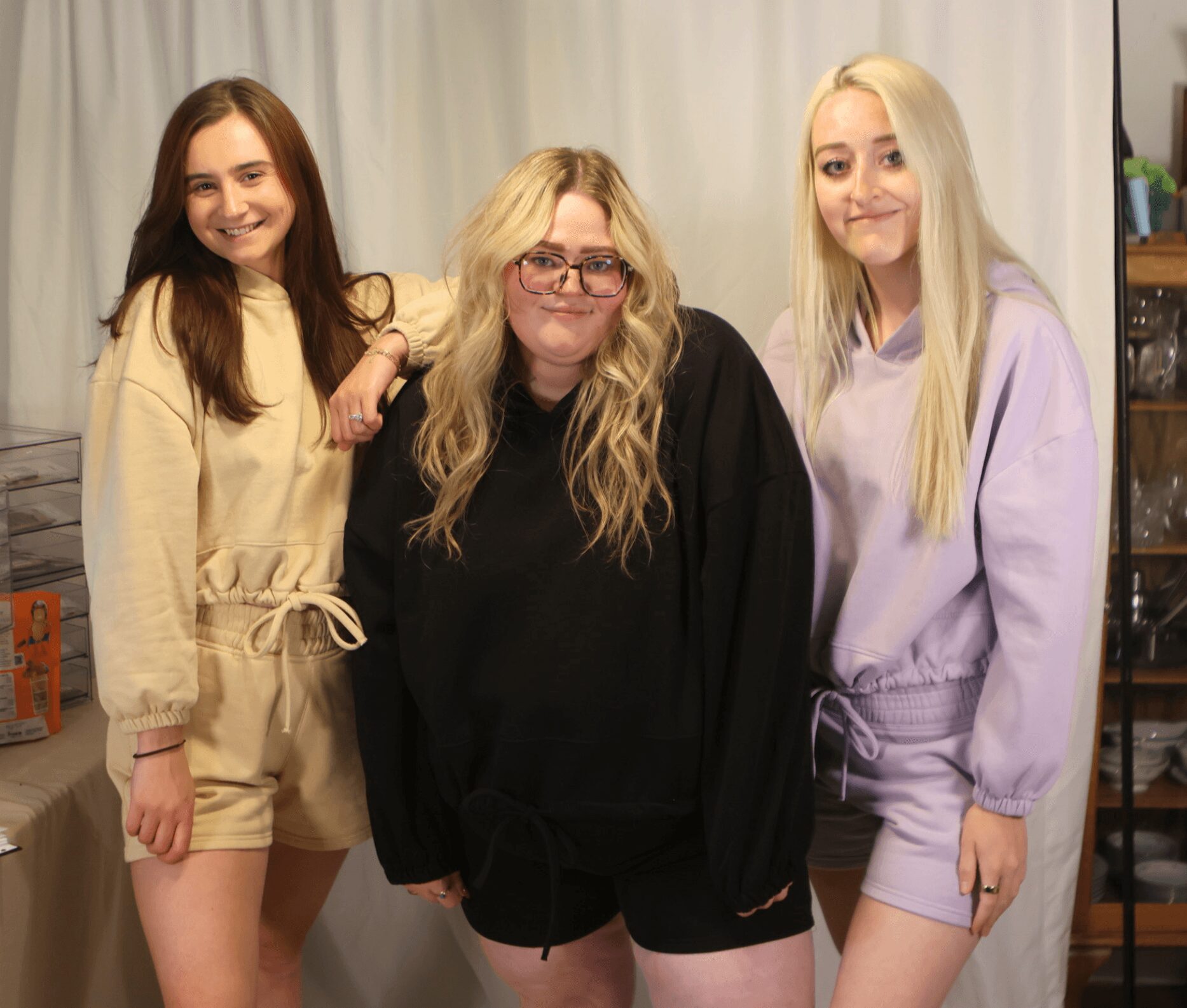 Three women standing together indoors, smiling, with a curtain background and shelves on the right side.