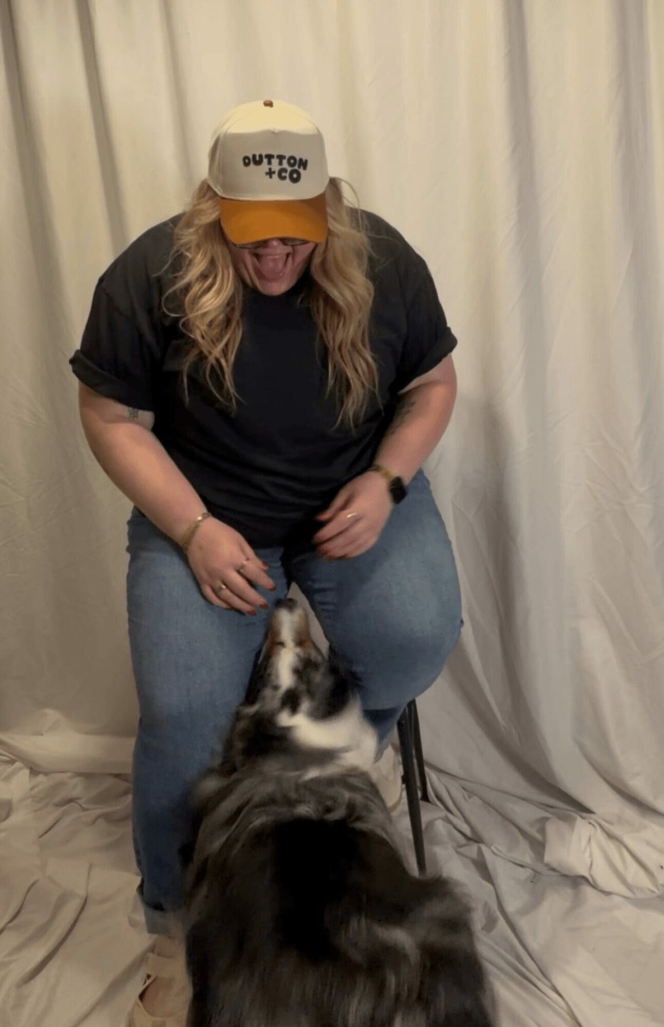 Person with long hair wearing a cap and black shirt interacts with a black and white dog indoors, with curtains in background.