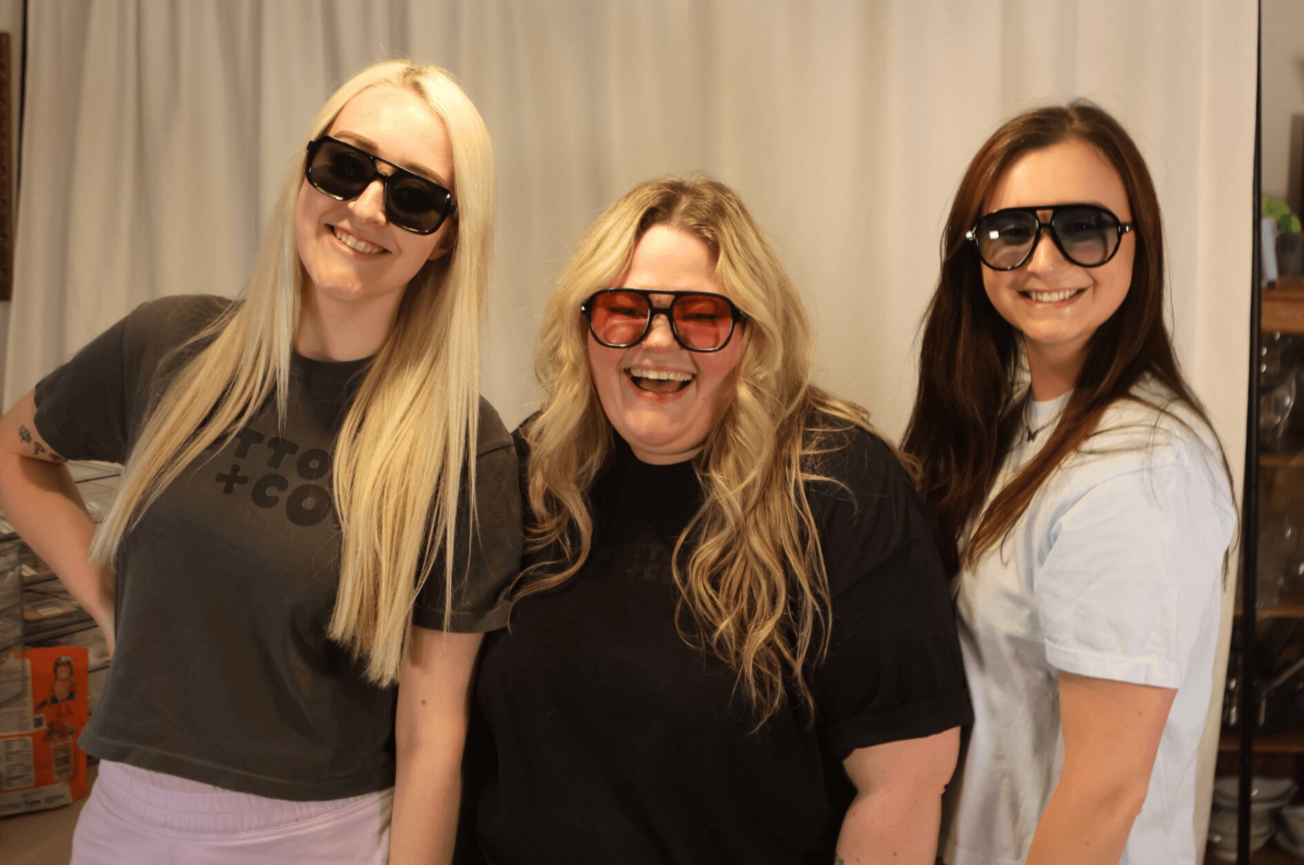 Three women smiling, wearing sunglasses, standing indoors in front of a curtain, with light-colored hair and casual clothing.