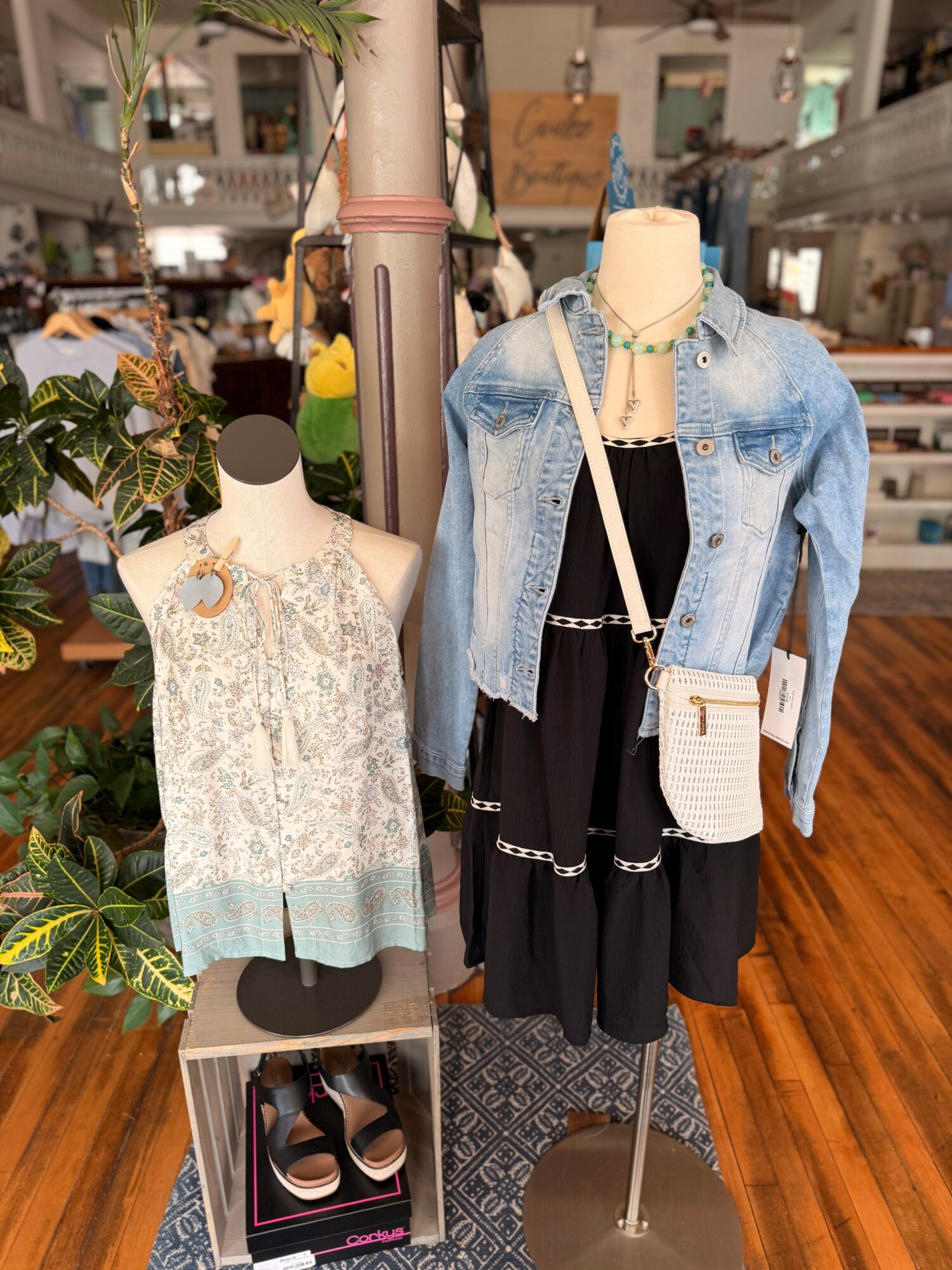 Mannequin dressed in a black dress with a denim jacket, white purse, and necklace, displayed in a store with plants and wooden floors.