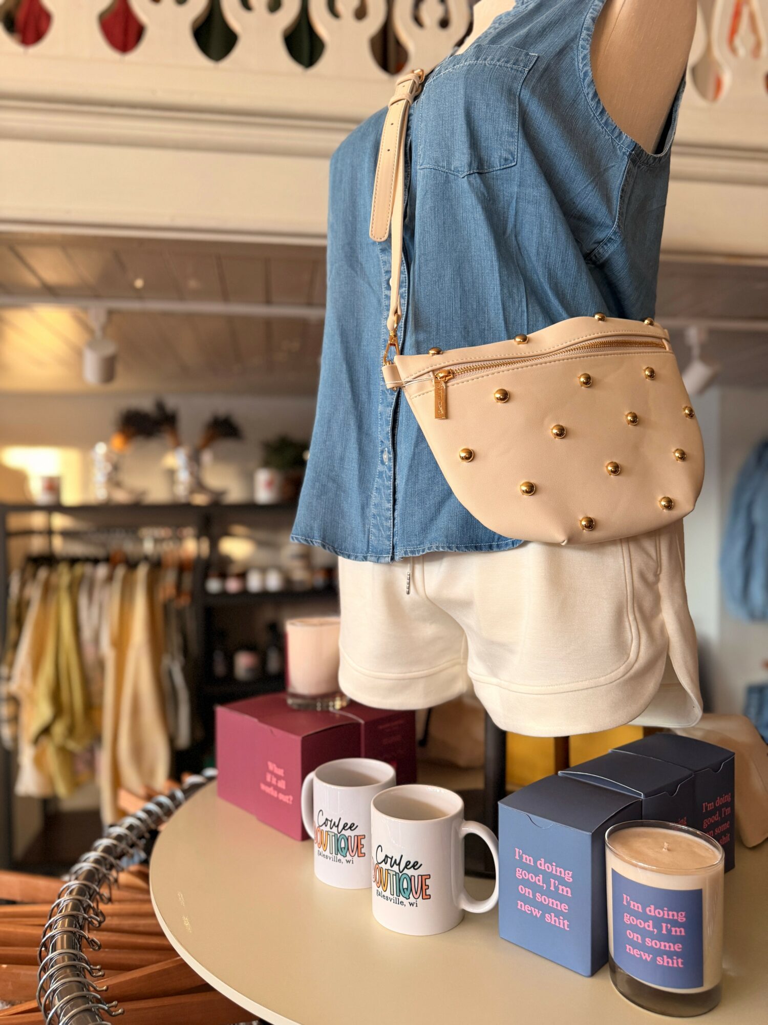 Person wearing a blue top and white shorts with a beige bag, standing near a table with mugs and candles.