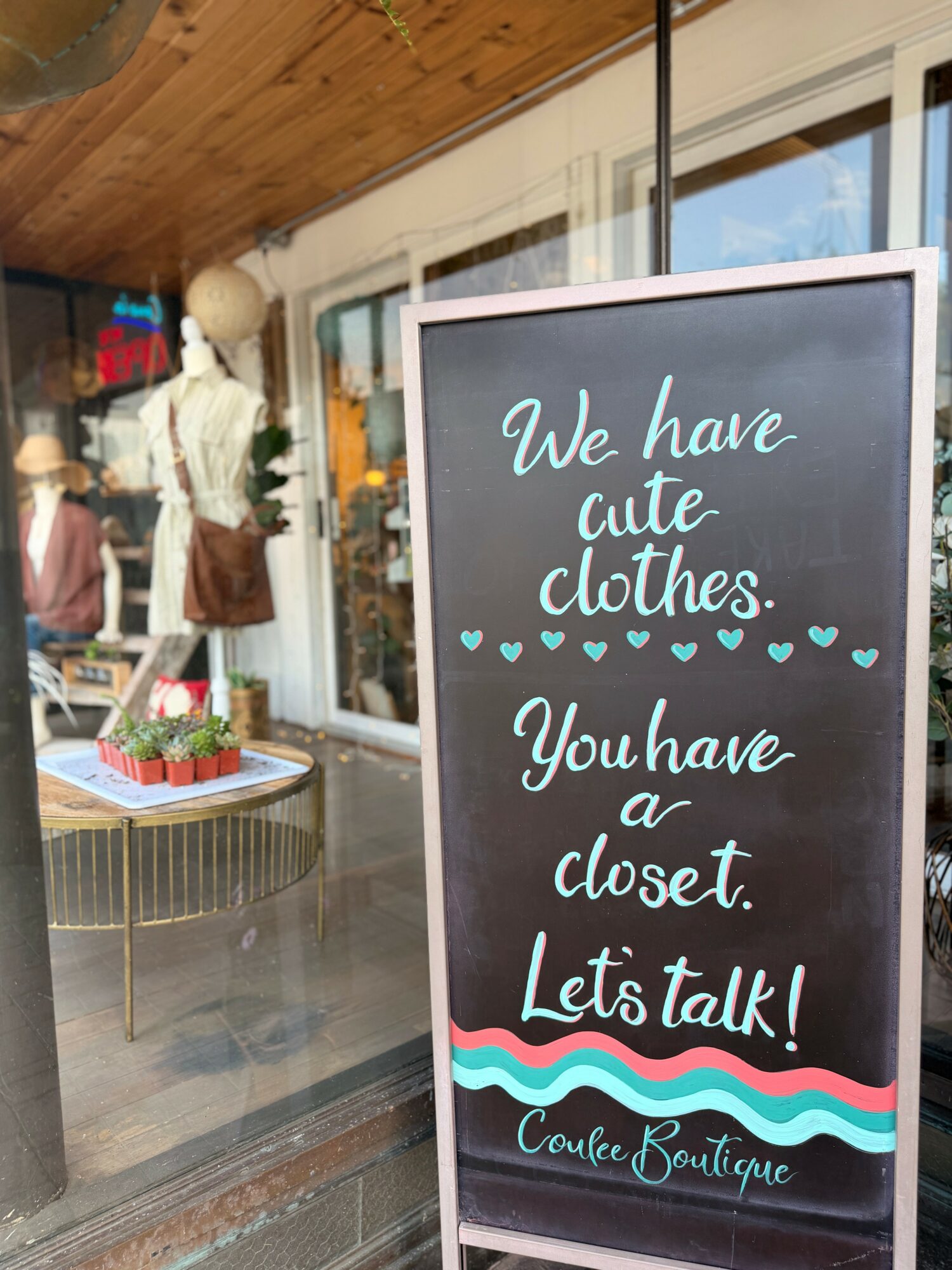 Store window display with mannequins and a chalkboard sign outside, advertising clothing and a closet, with a wooden ceiling.