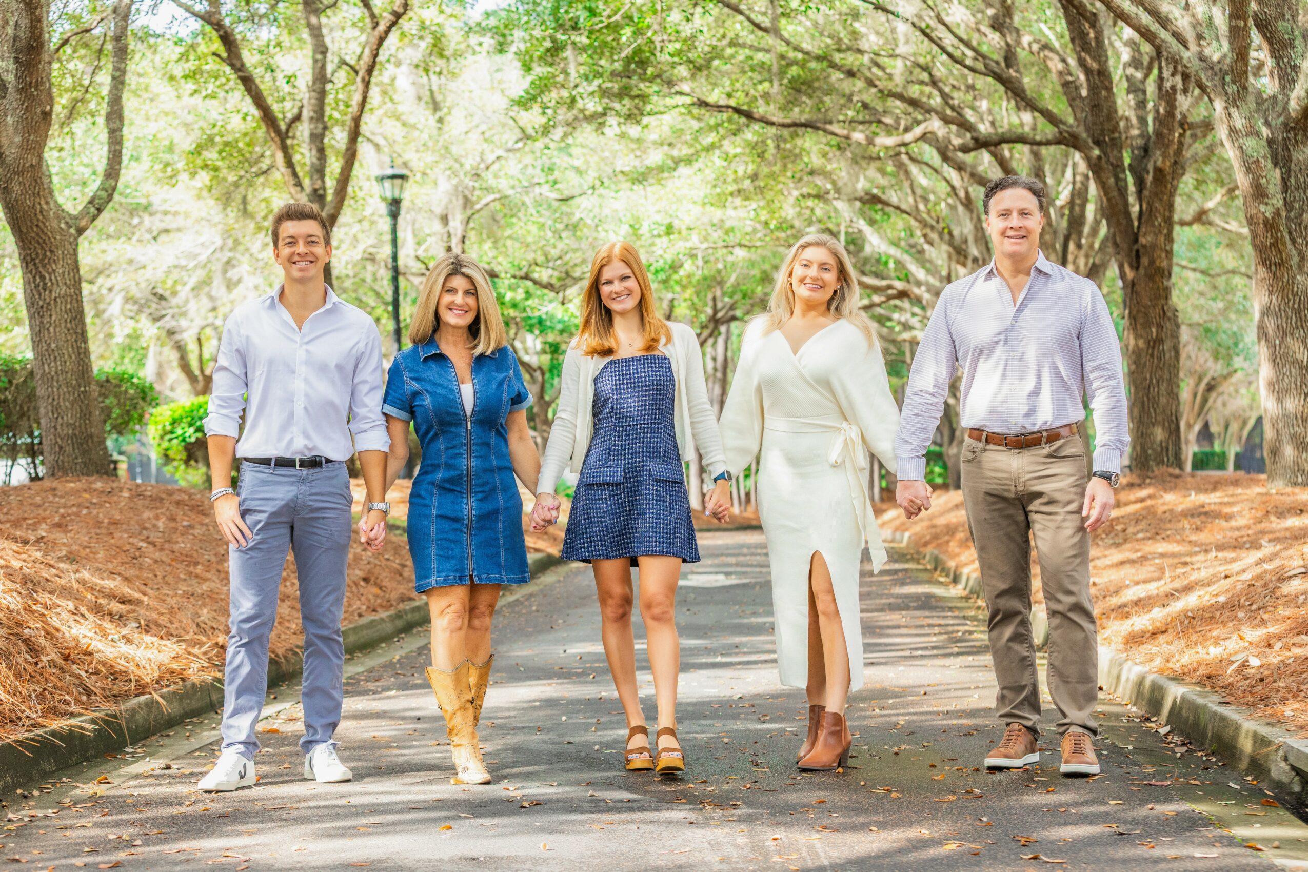 Group of five people walking on a park path holding hands, surrounded by trees and greenery.