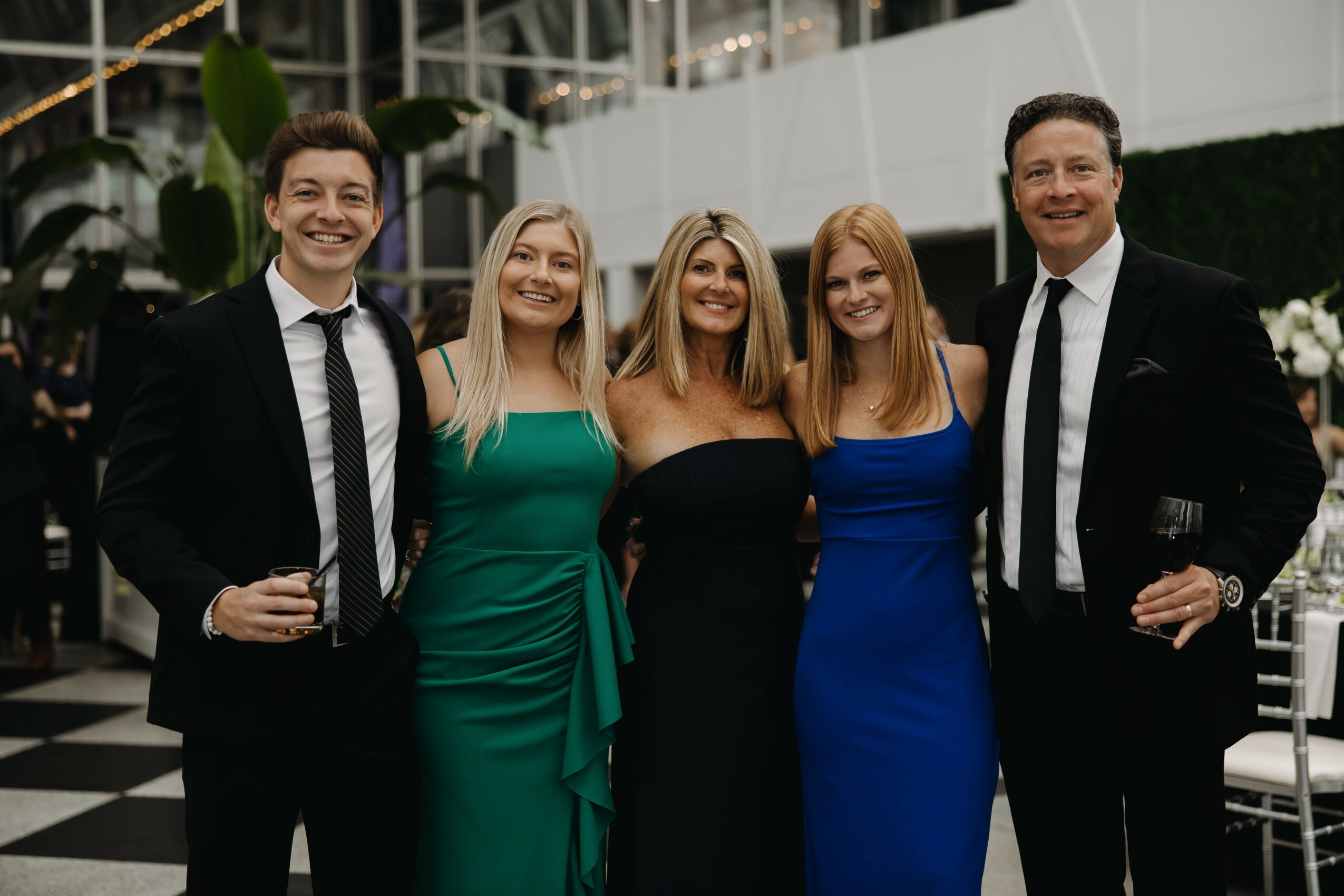 Group of five people in formal attire smiling indoors, three women in dresses and two men in suits, holding drinks.