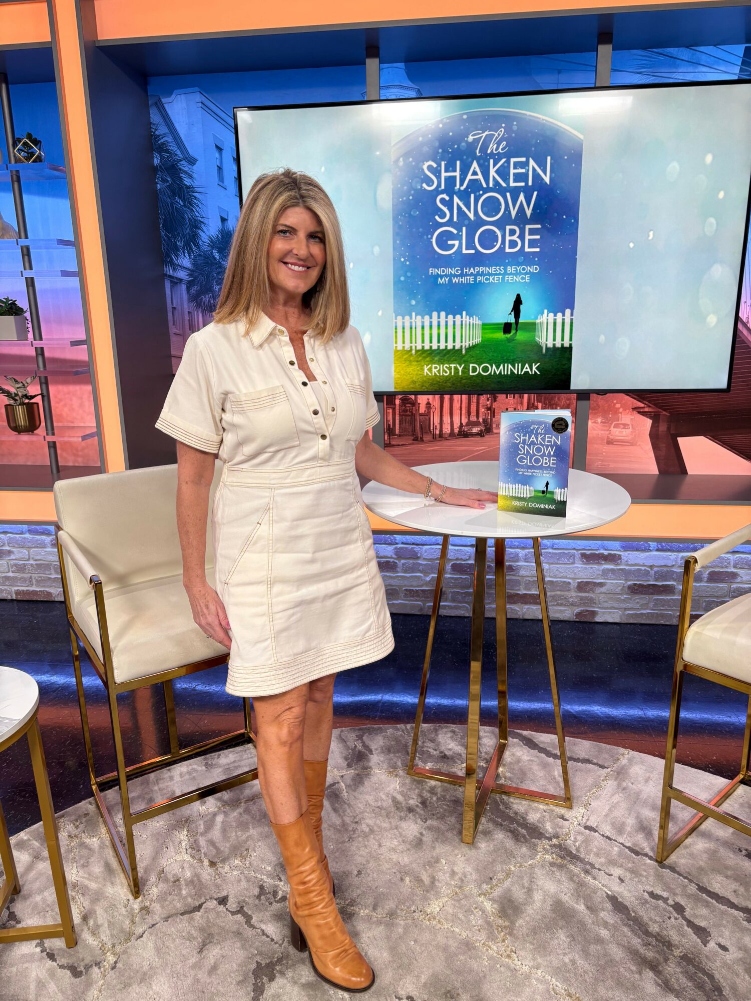 Woman in a white dress standing in front of a screen displaying a book cover, with a chair and table nearby.