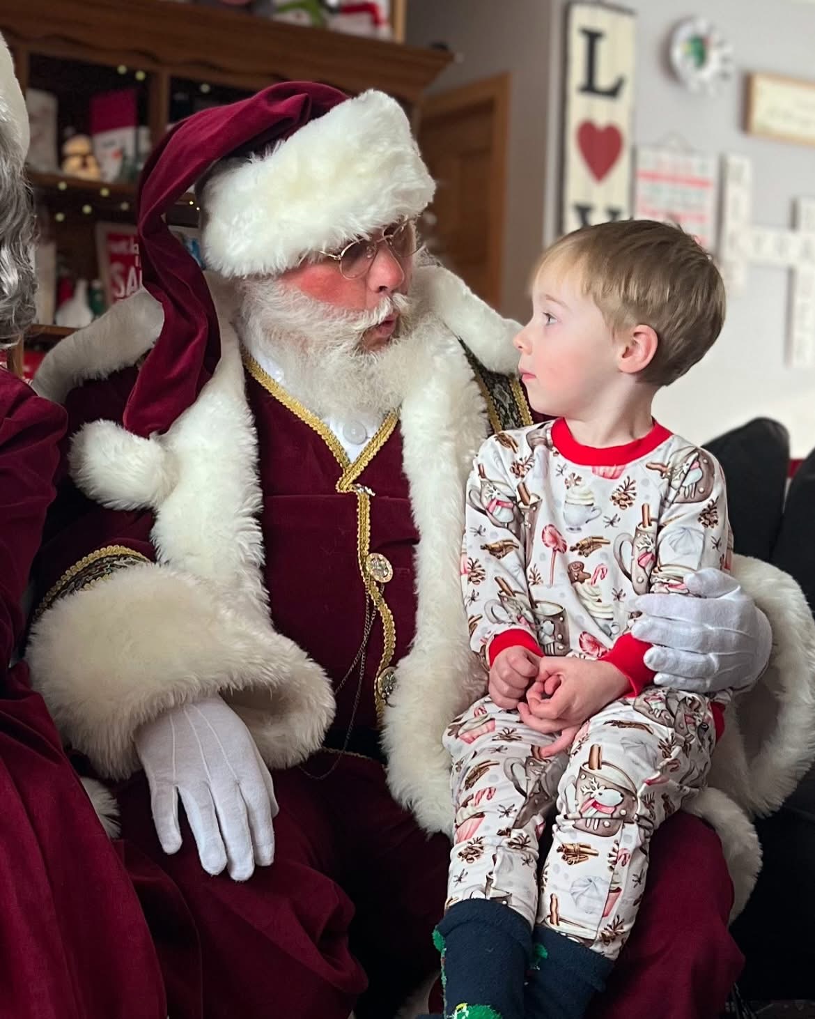 Santa Claus in a velvet suit and white gloves sits with a young boy in festive pajamas, indoors with holiday decorations.