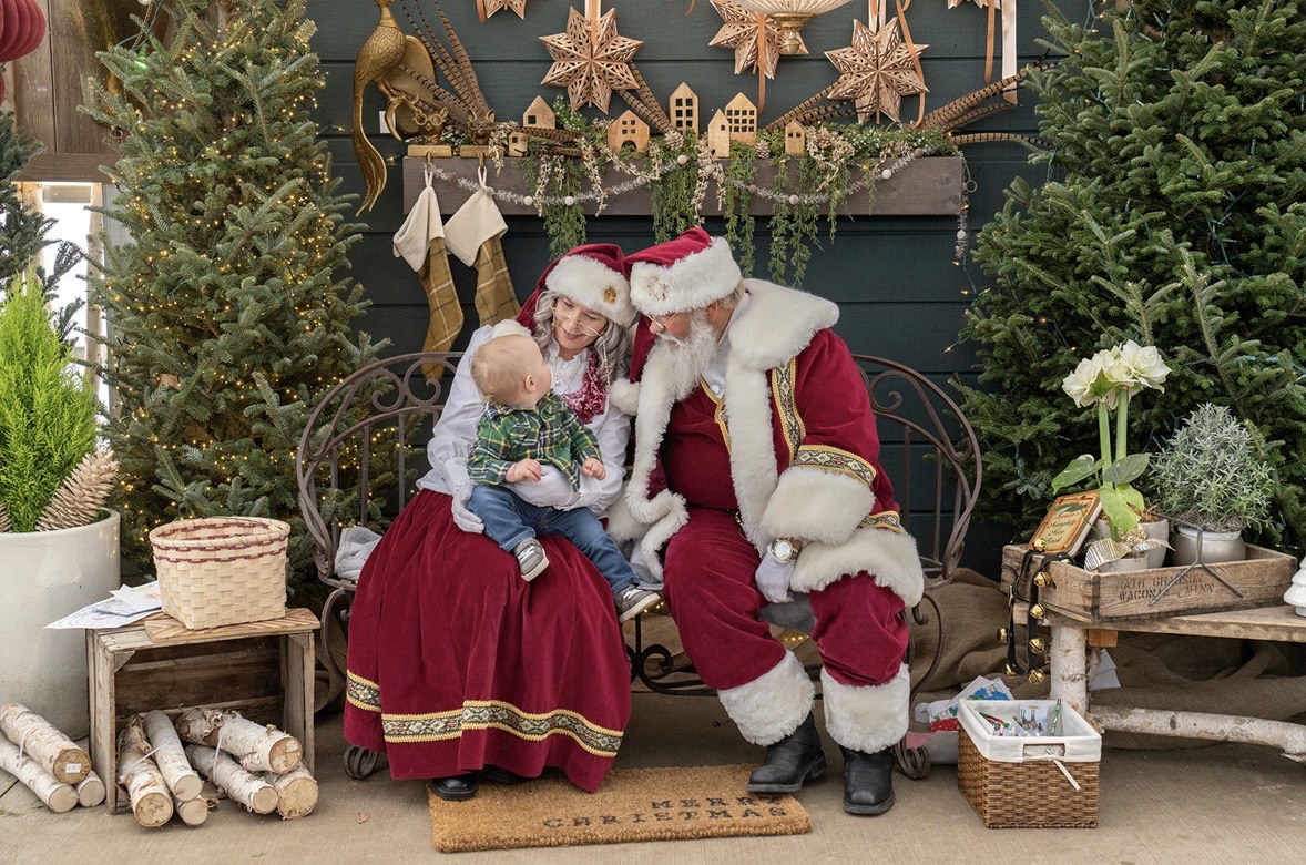 Child sitting on Santa Claus's lap, both wearing Santa hats, surrounded by Christmas trees and decorations.