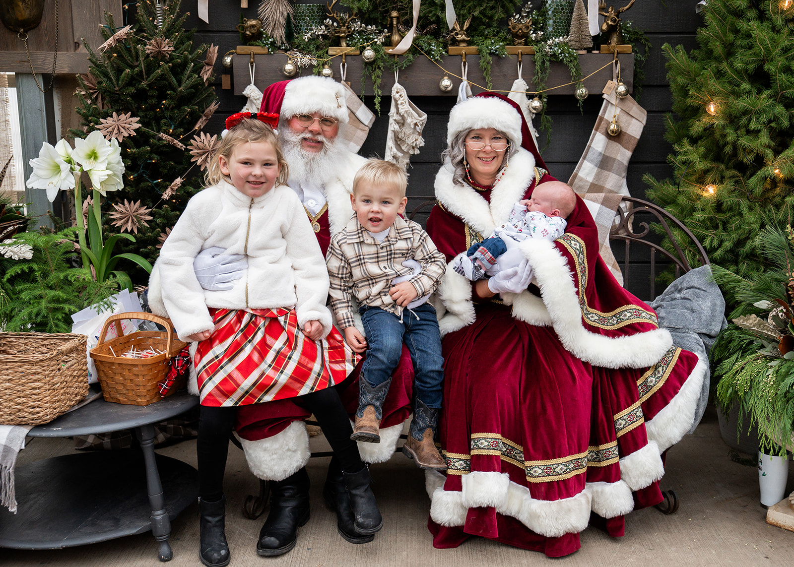 Children and adults in Christmas costumes sit with Santa Claus and Mrs. Claus, surrounded by decorated Christmas trees and plants.