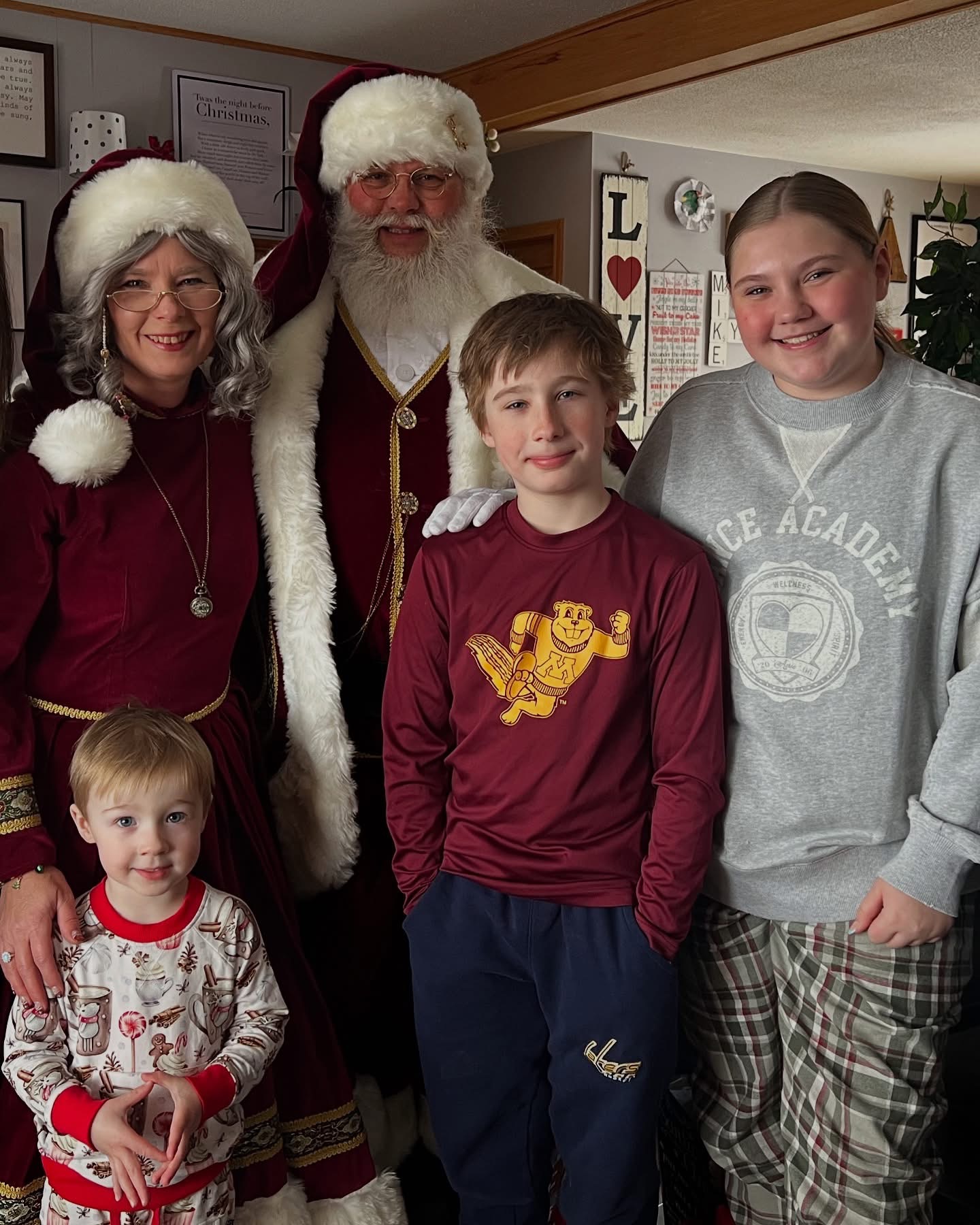 Group of five people, including two children, posing indoors with Christmas decorations, smiling, with a Santa figure in the background.