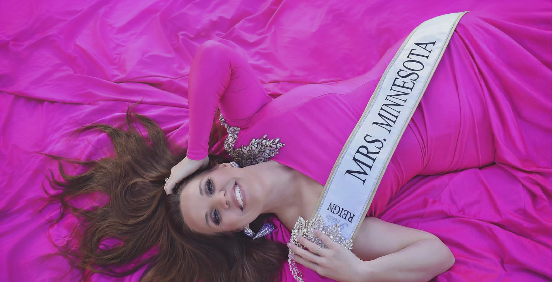 Woman lying on pink fabric wearing a sash that reads 'Mrs. Minnesota', holding a crown, with long hair spread out.