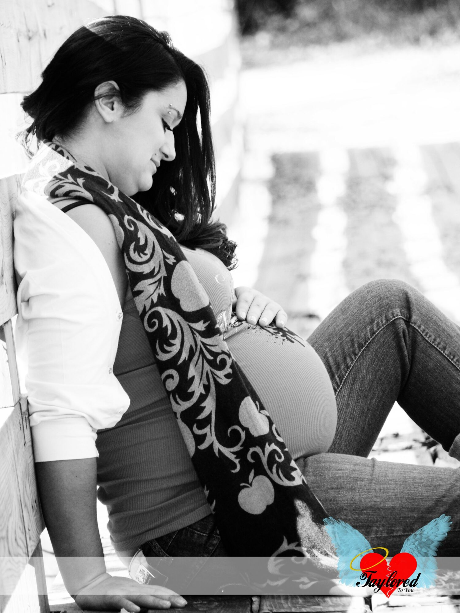 Pregnant woman sitting outdoors, looking down, with long dark hair, wearing a patterned scarf, white shirt, and jeans.