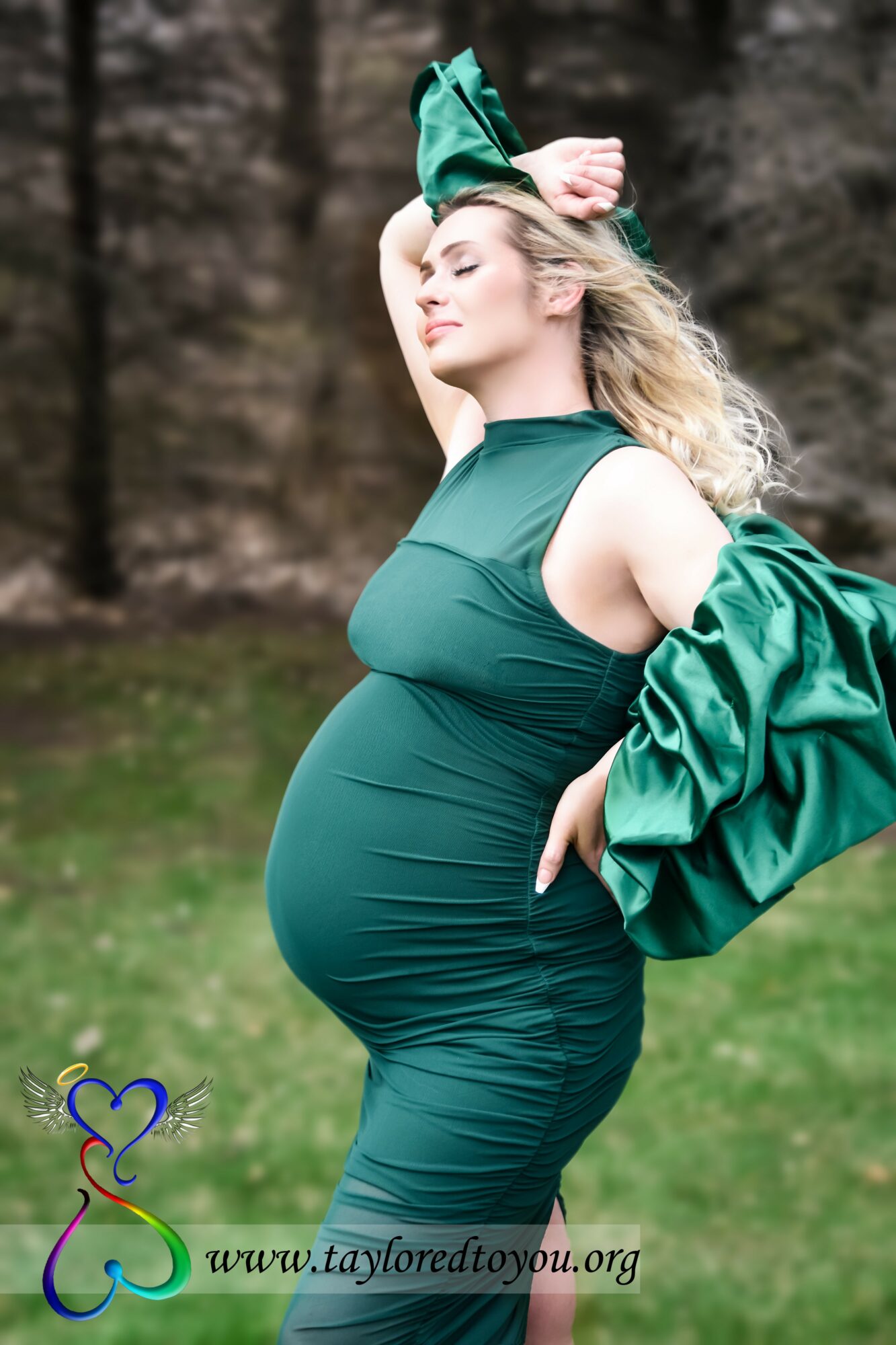 Pregnant woman in a green dress outdoors, with her eyes closed and one arm raised, holding a jacket.