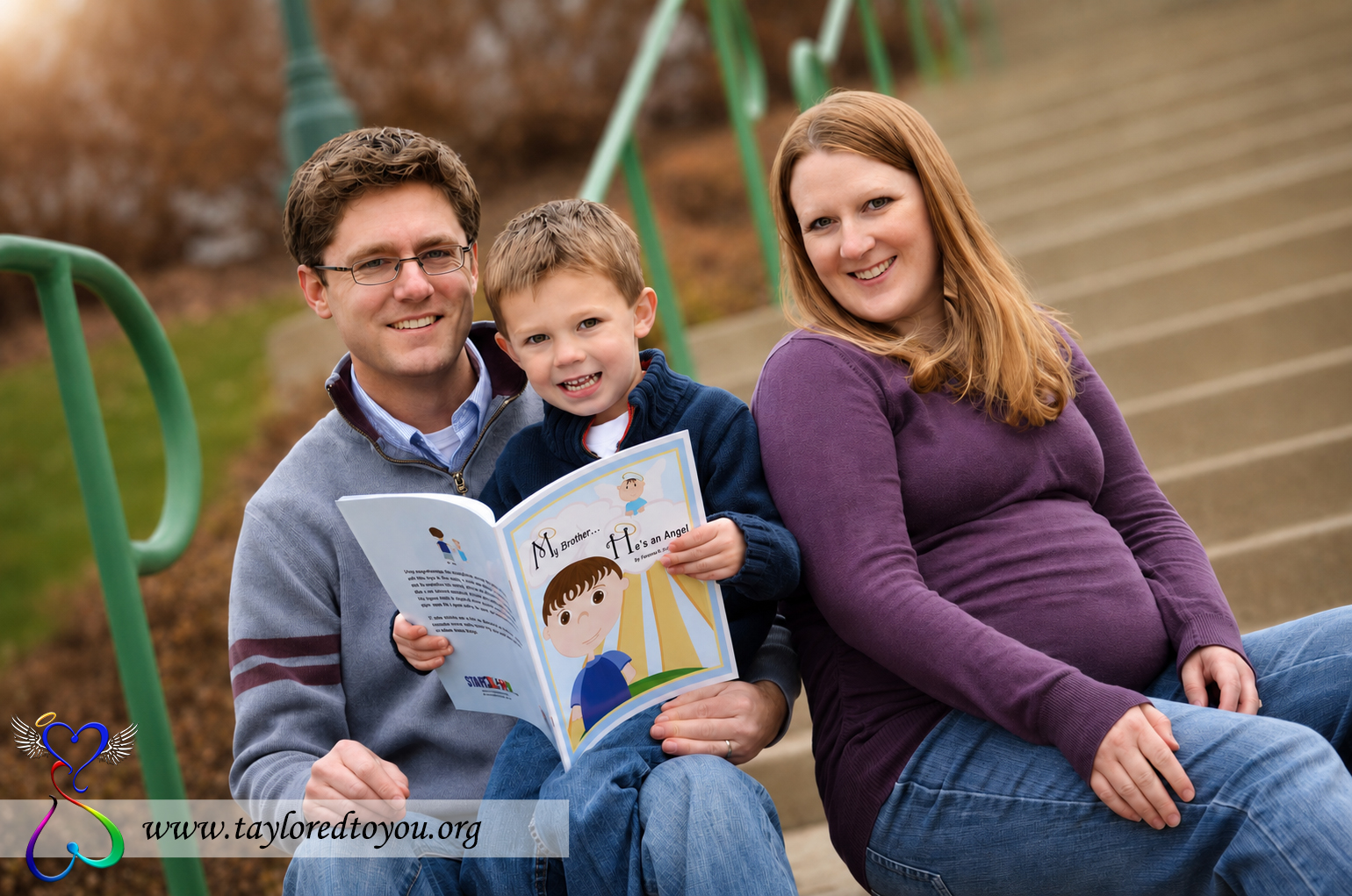 Family of three sitting on outdoor stairs, reading a book together, smiling, with green railings in background.