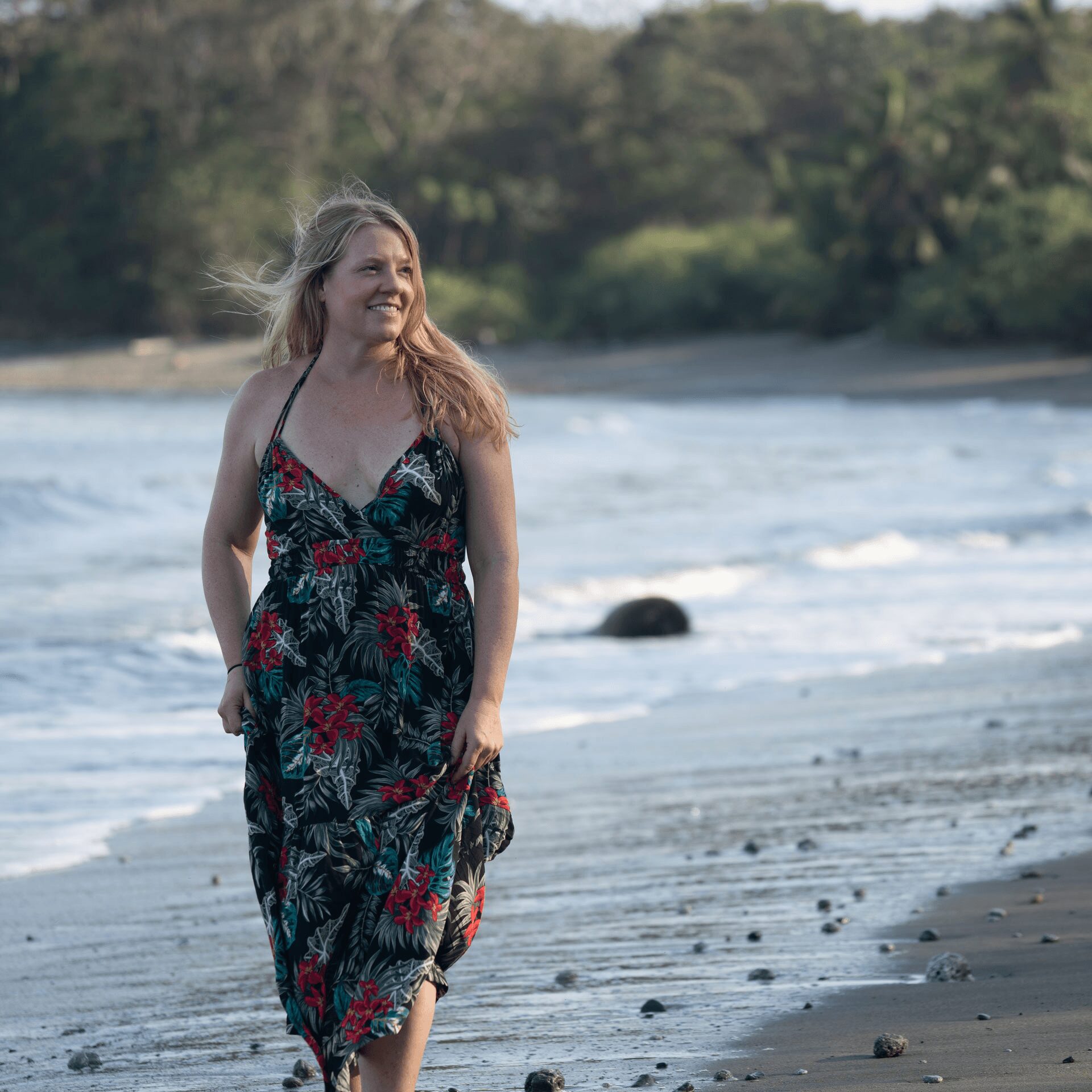 Woman in a floral dress walking on a beach with water and trees in the background.