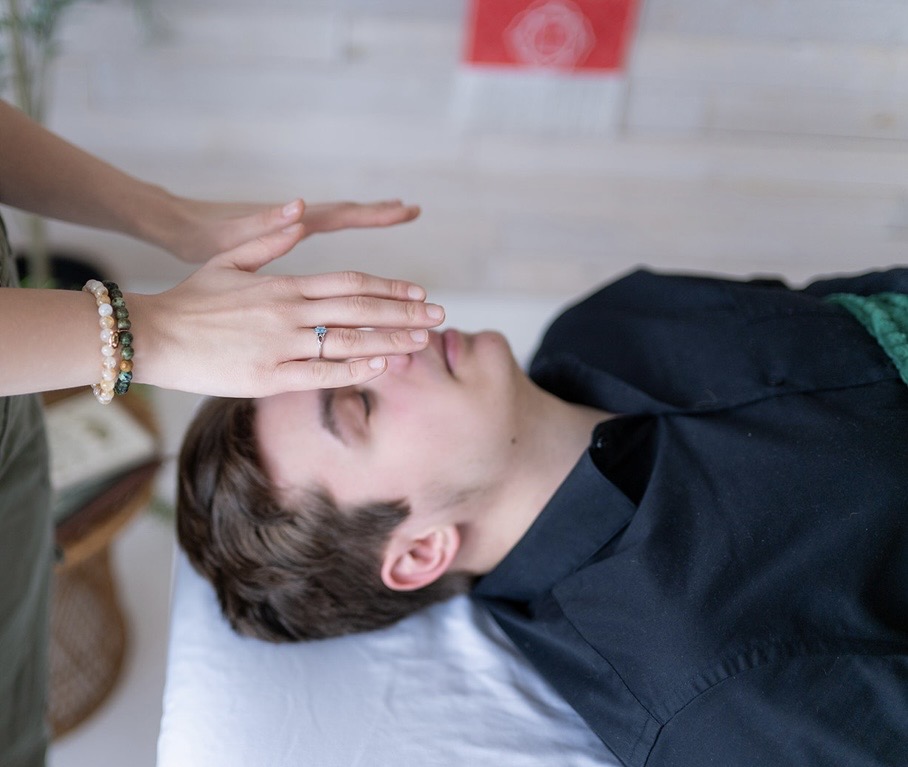 Person lying down with eyes closed, receiving a forehead massage from another person with hands visible, in a room with a white wall.