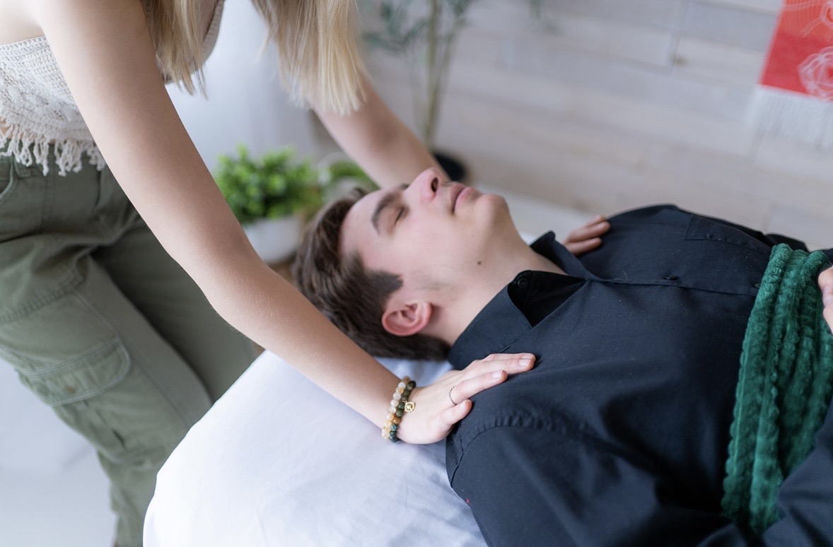 Person lying on a bed with eyes closed, receiving a massage or treatment from another person. The setting appears to be indoors.